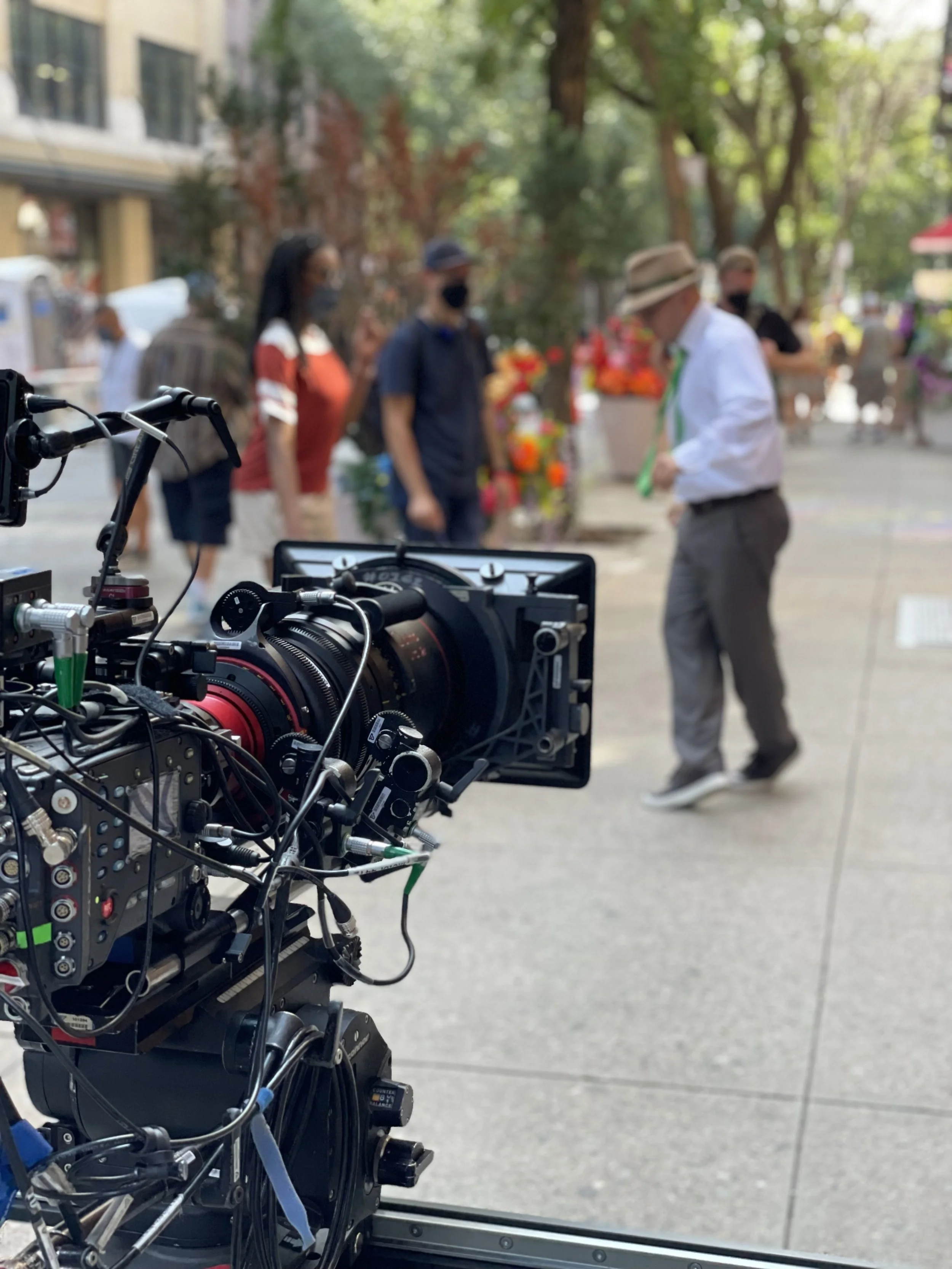 Close-up of a professional film cameraon a tripod, focused on a street scene with production crew.