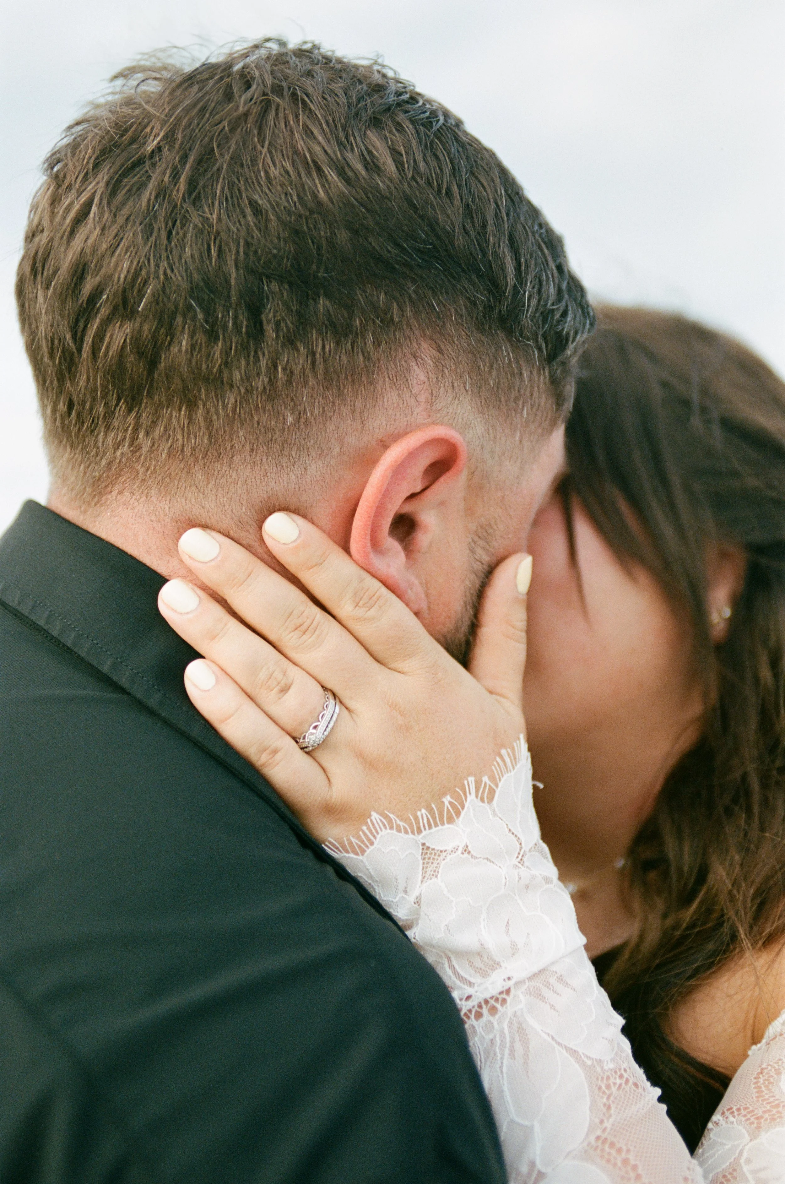 A man wearing a dark suit holding a woman in a white lace dress, with her hand on his face. The man has short, styled hair. The woman has long brown hair.