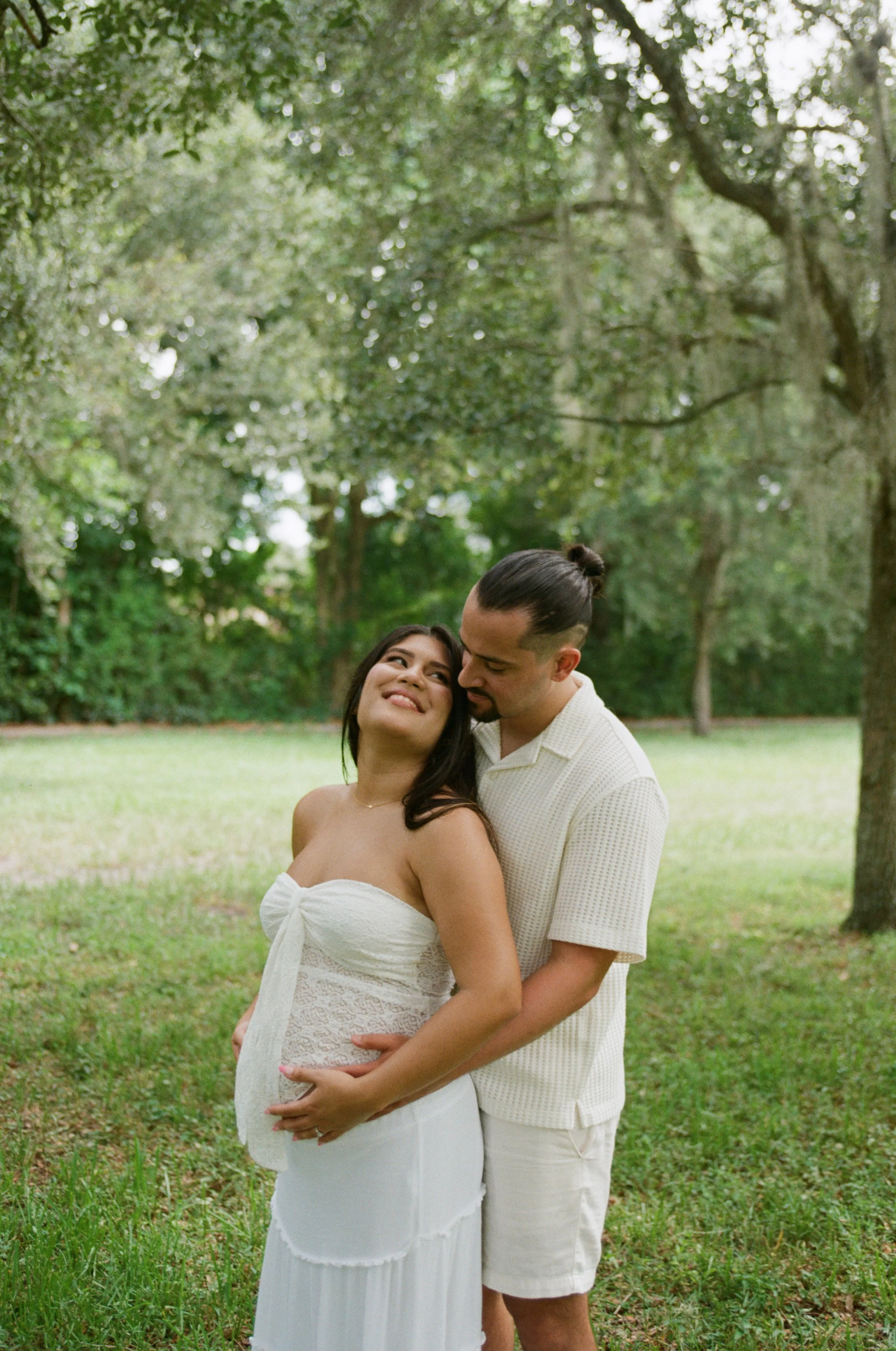 A couple standing outdoors in a park with green grass and trees, the woman in a strapless white dress and the man in a light-colored shirt and shorts, embracing affectionately.