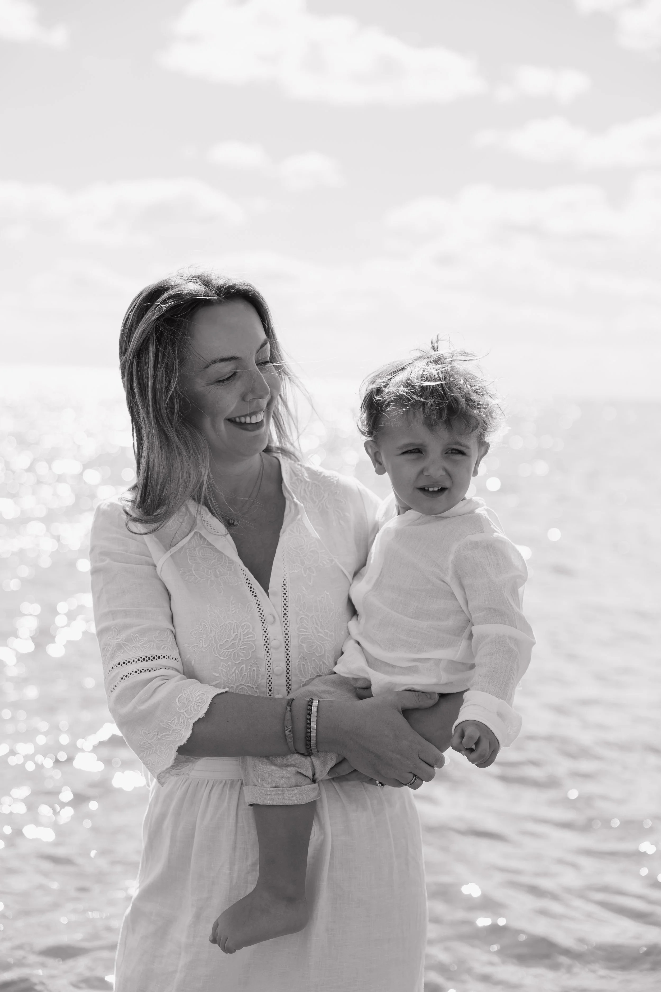 A woman holding a young child by the beach, smiling and looking to the side, with the ocean and clouds in the background.