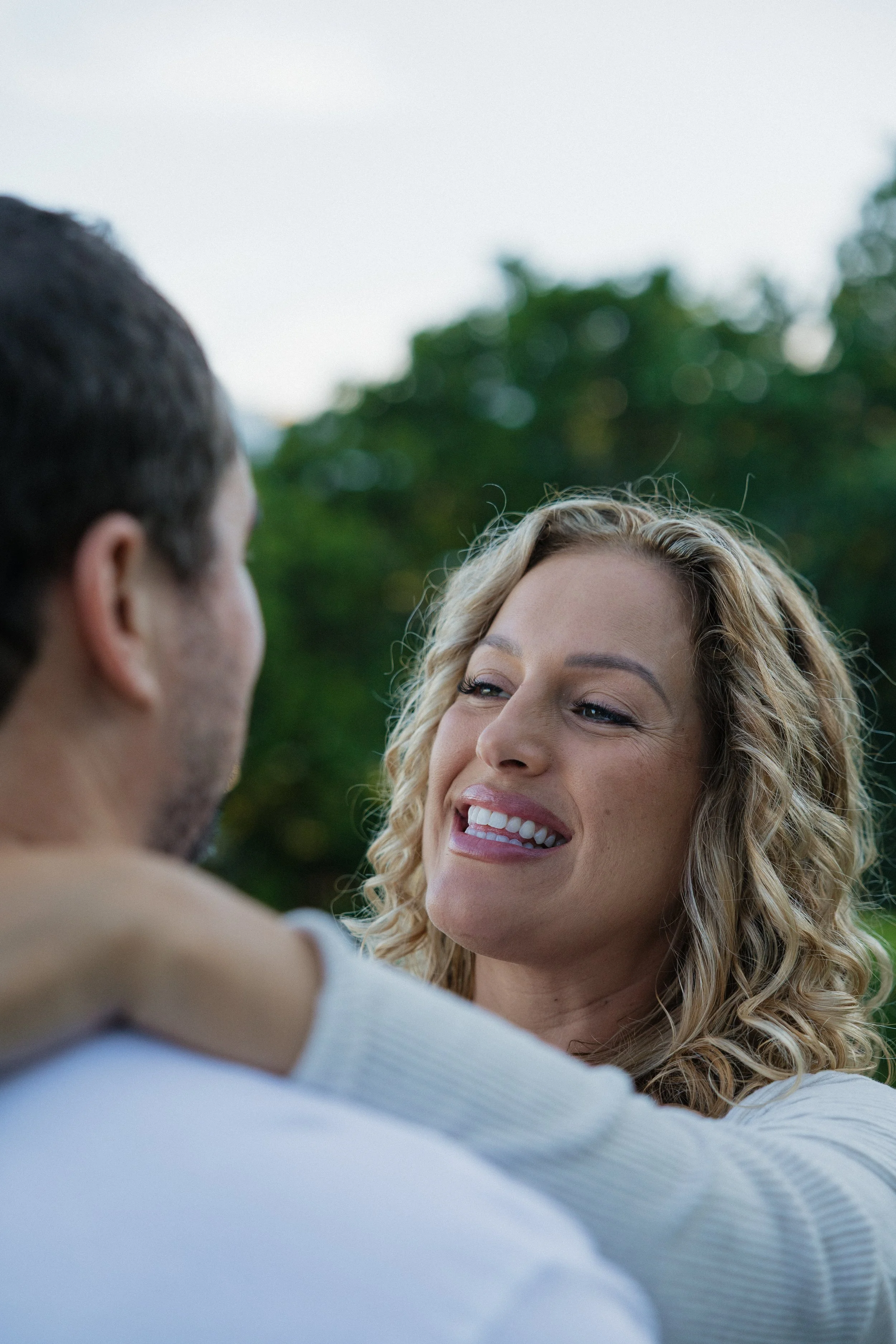 A woman with long curly blonde hair smiling at a man with dark hair, both outdoors with blurred green trees in the background.