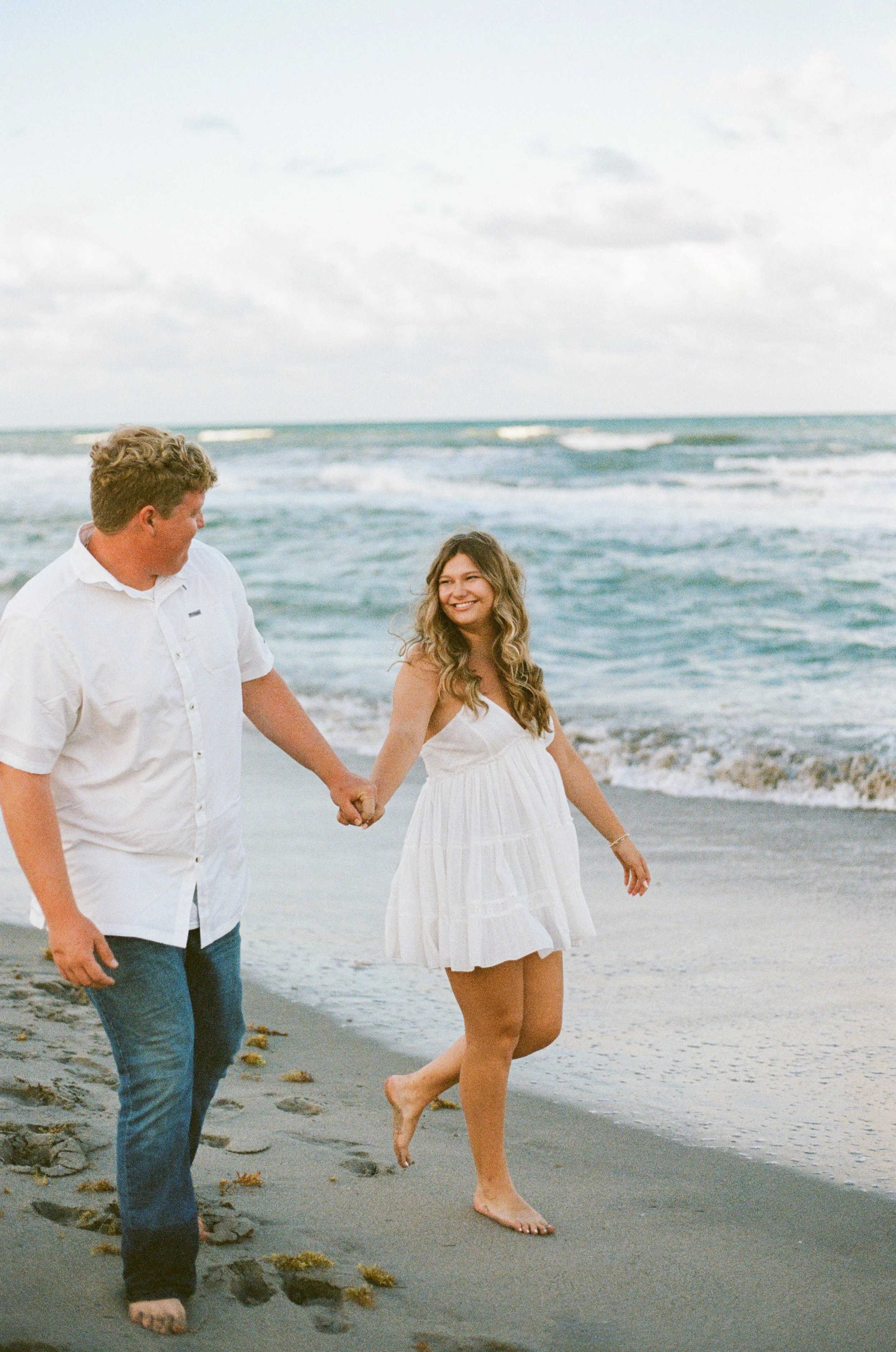 A couple walking hand in hand on the beach, with the woman smiling and looking at the man. The man is wearing a white button-up shirt and jeans, and the woman is in a white dress. The ocean waves are in the background.