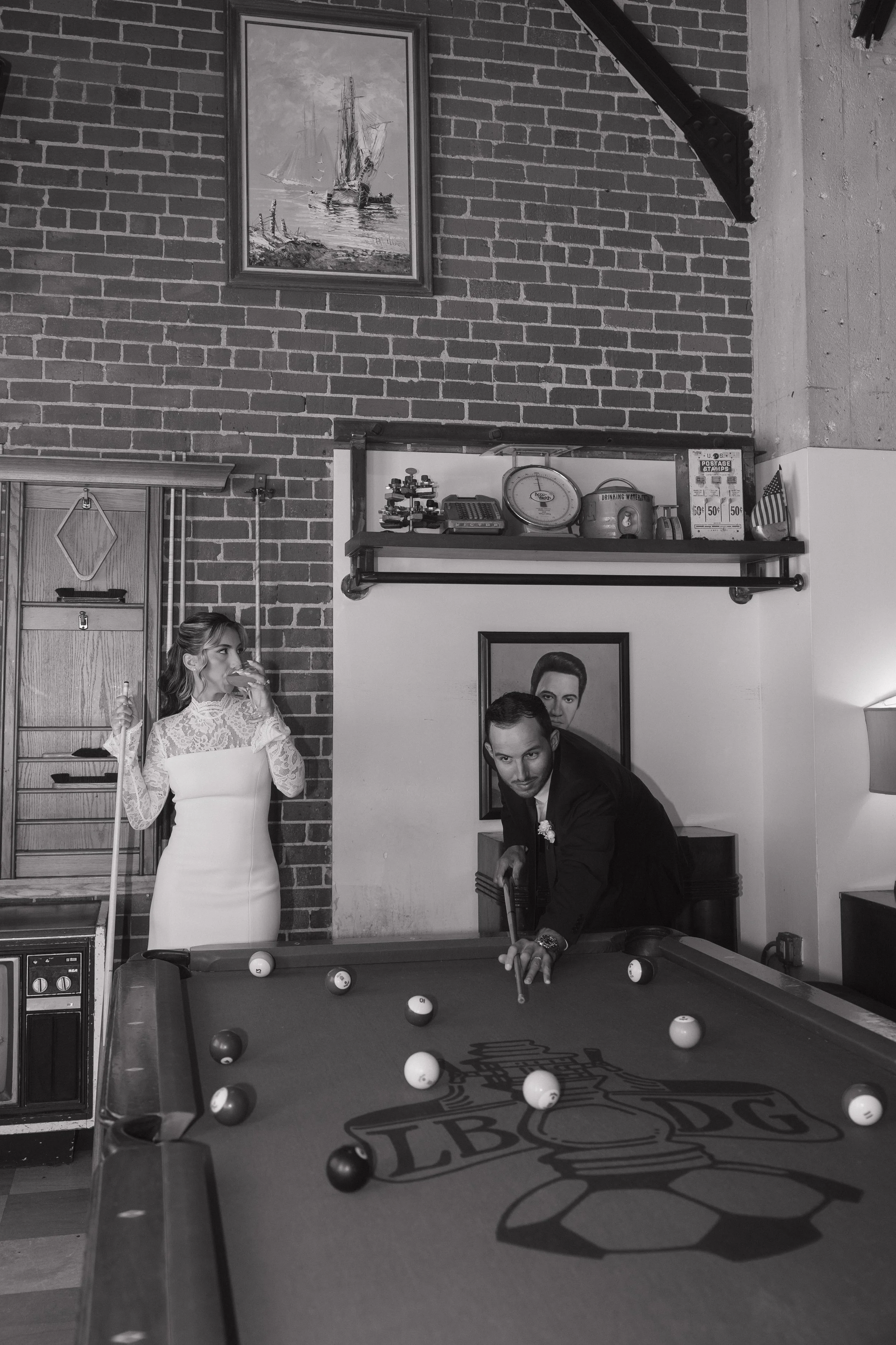 People playing pool in a bar with brick walls, a framed painting of ships, and shelves with various items. One woman in a white lace dress is watching while a man in a dark suit takes his shot.