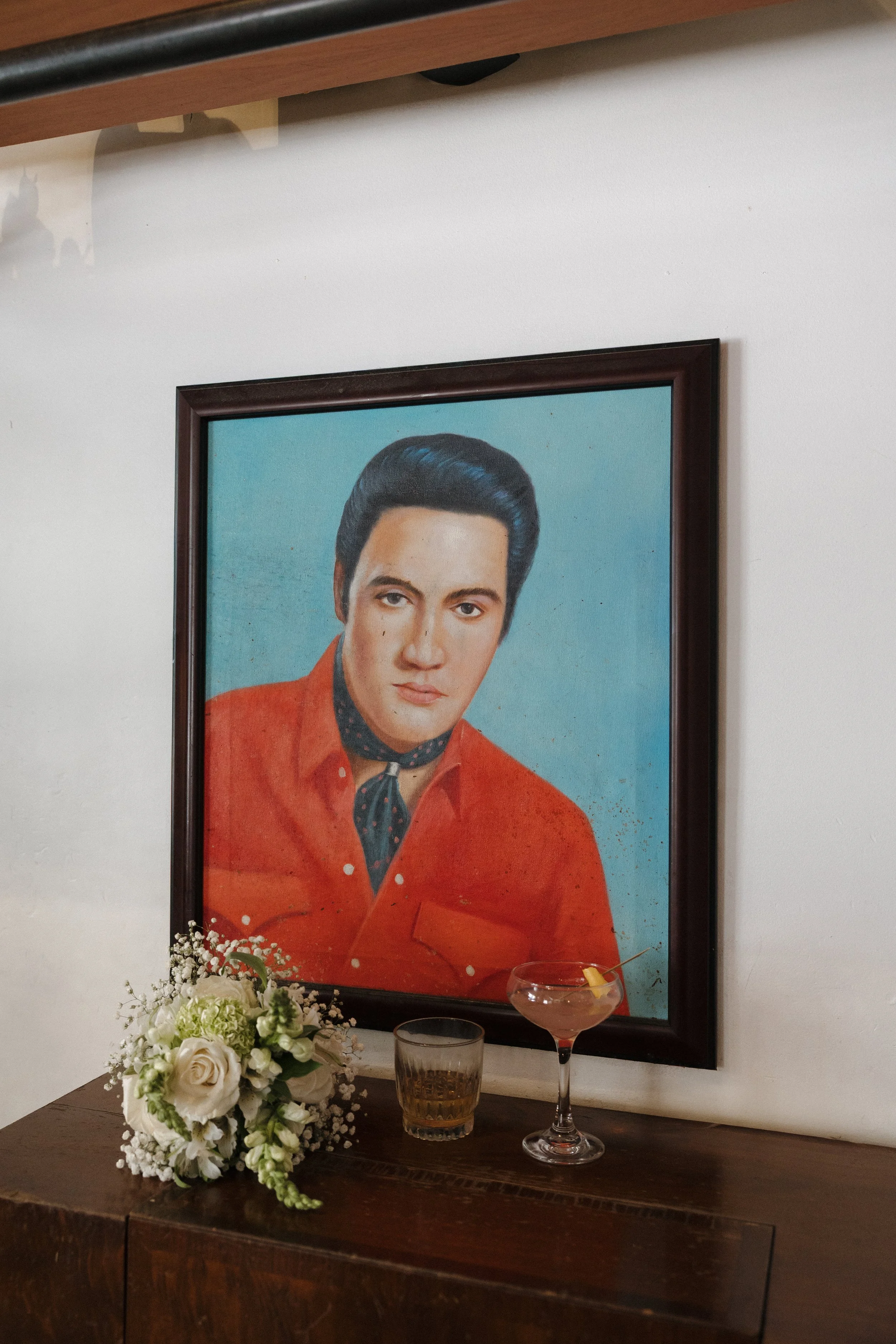 A framed picture of Elvis Presley hanging on a white wall with a floral arrangement and drinks on a table below.