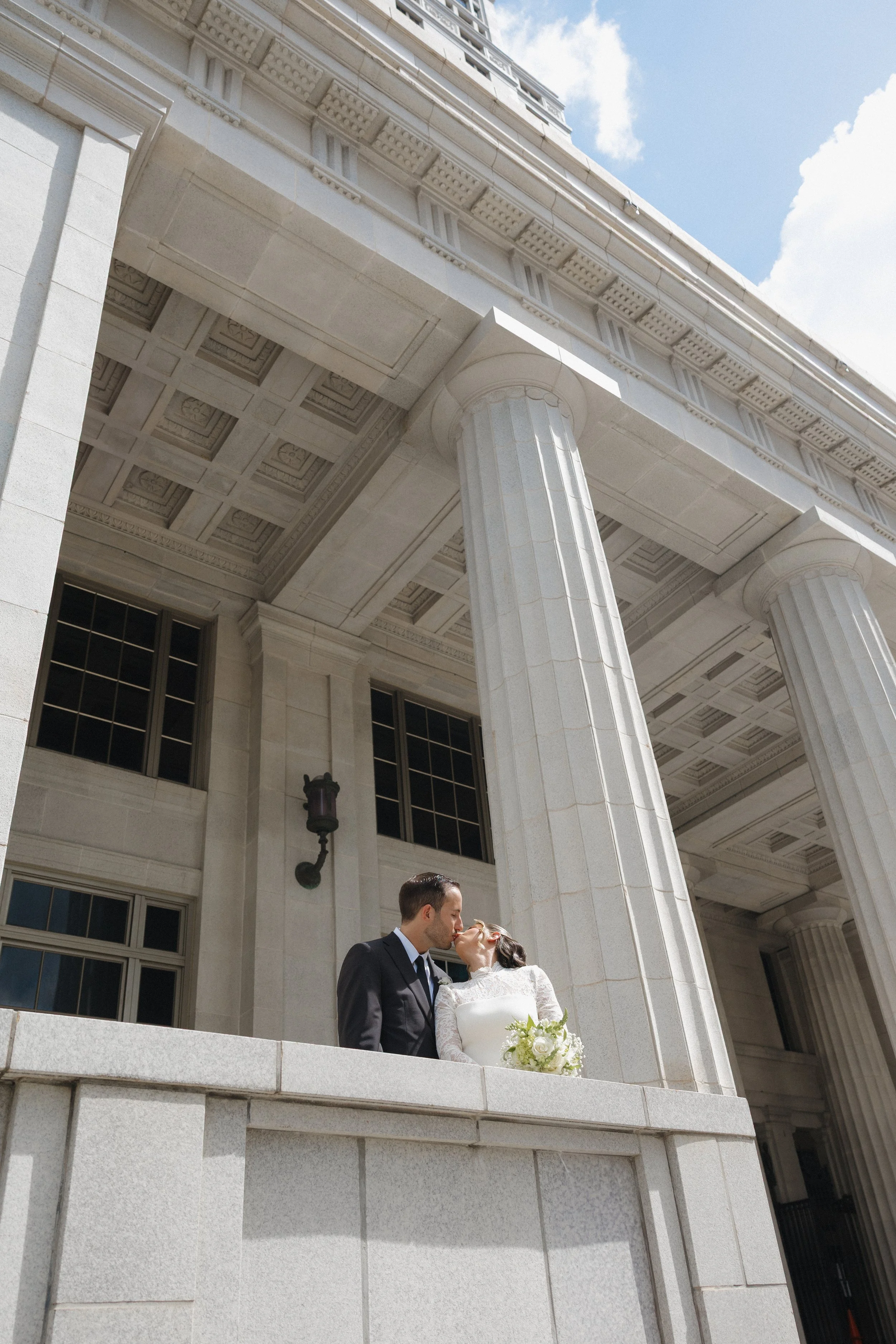 A newlywed couple kisses on the steps of a classical building with large columns and detailed architectural features under a partly cloudy sky.