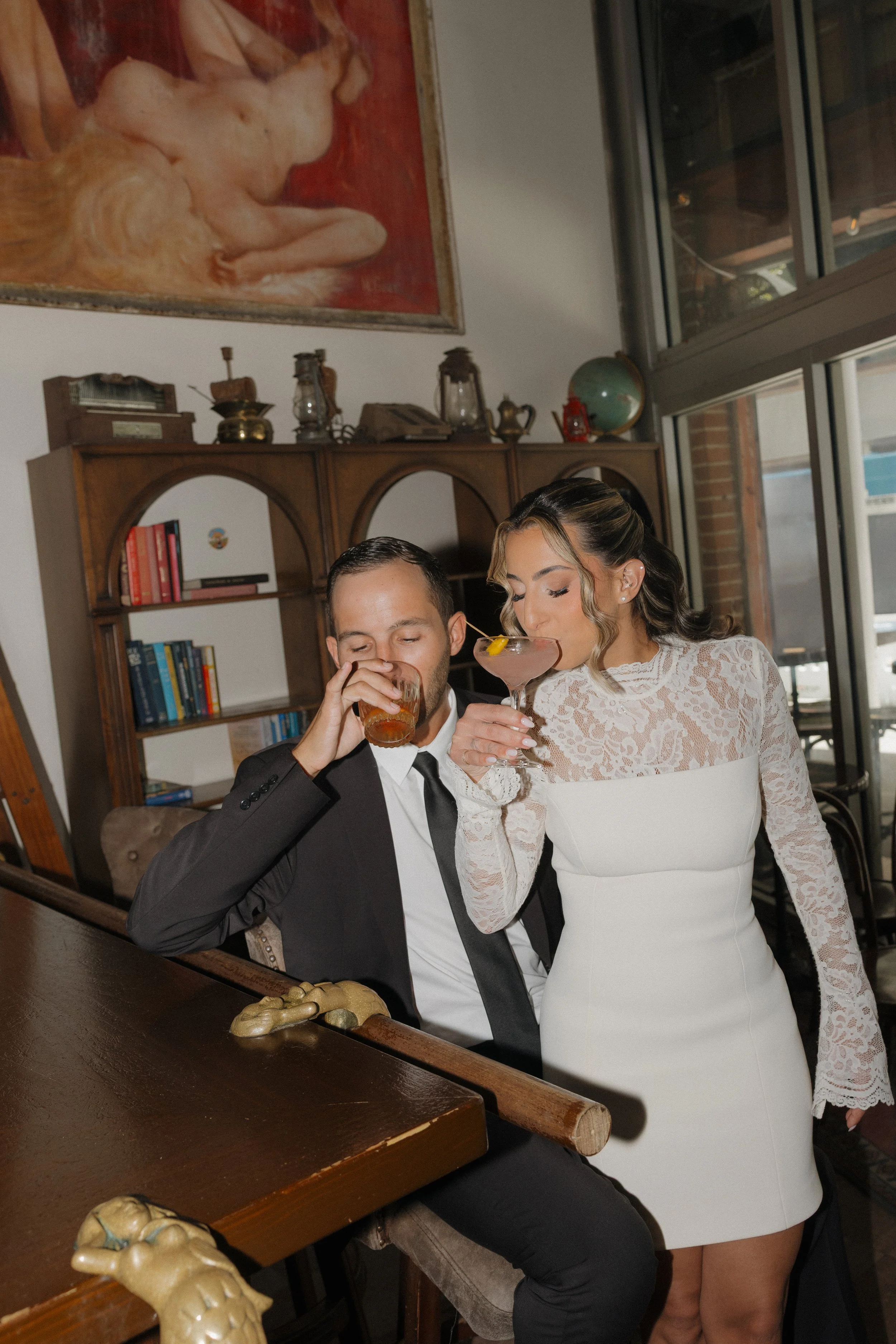 A man in a tuxedo and a woman in a white lace dress are enjoying drinks at a social event. The woman stands close to the man, who is seated and sipping from a glass. The setting appears to be indoors, decorated with vintage items and a large painting