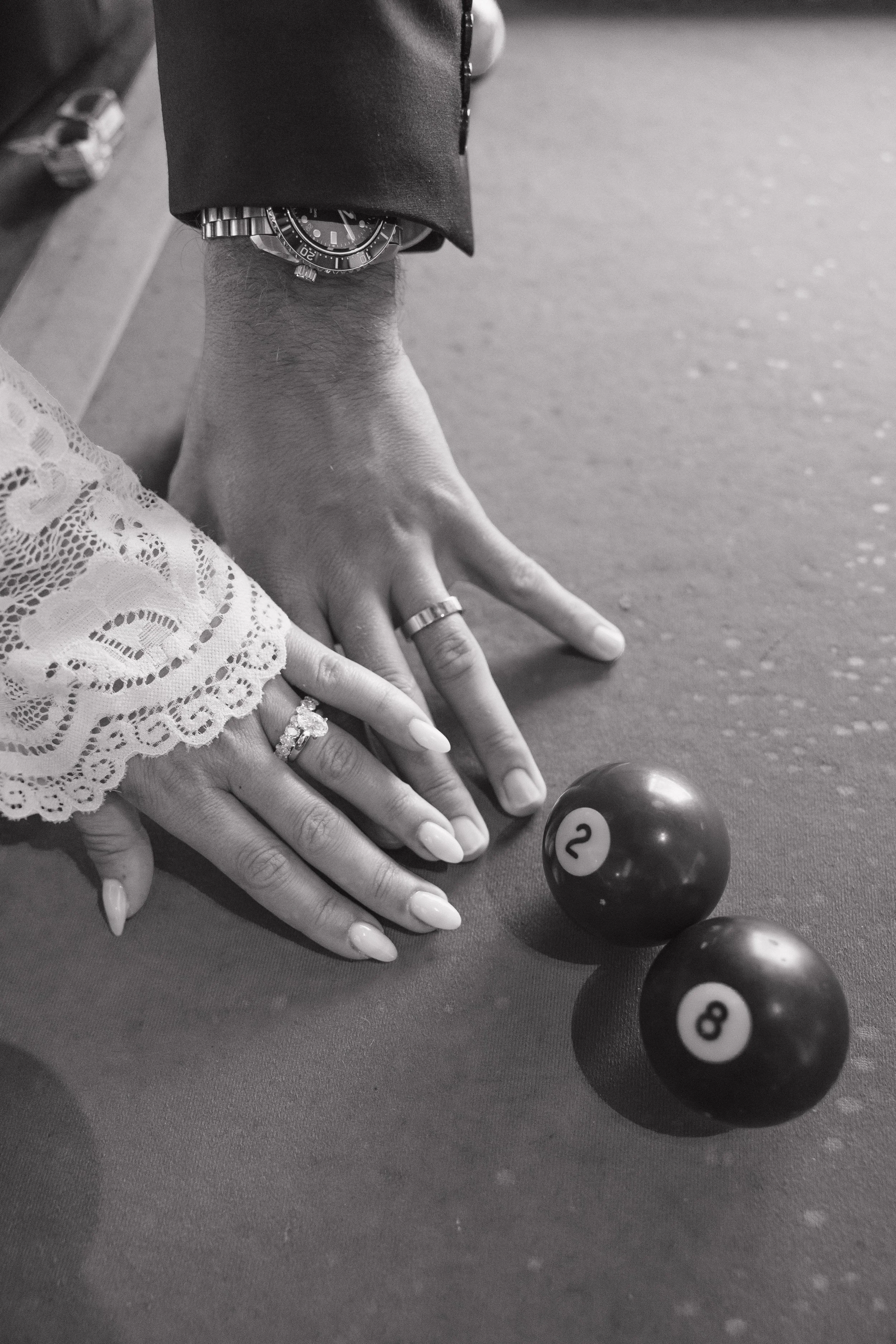 Close-up of a person's hand resting on a billiard table with a lace sleeve, wearing rings, with a wristwatch on the left wrist, near four billiard balls numbered 2, 8, and 4.