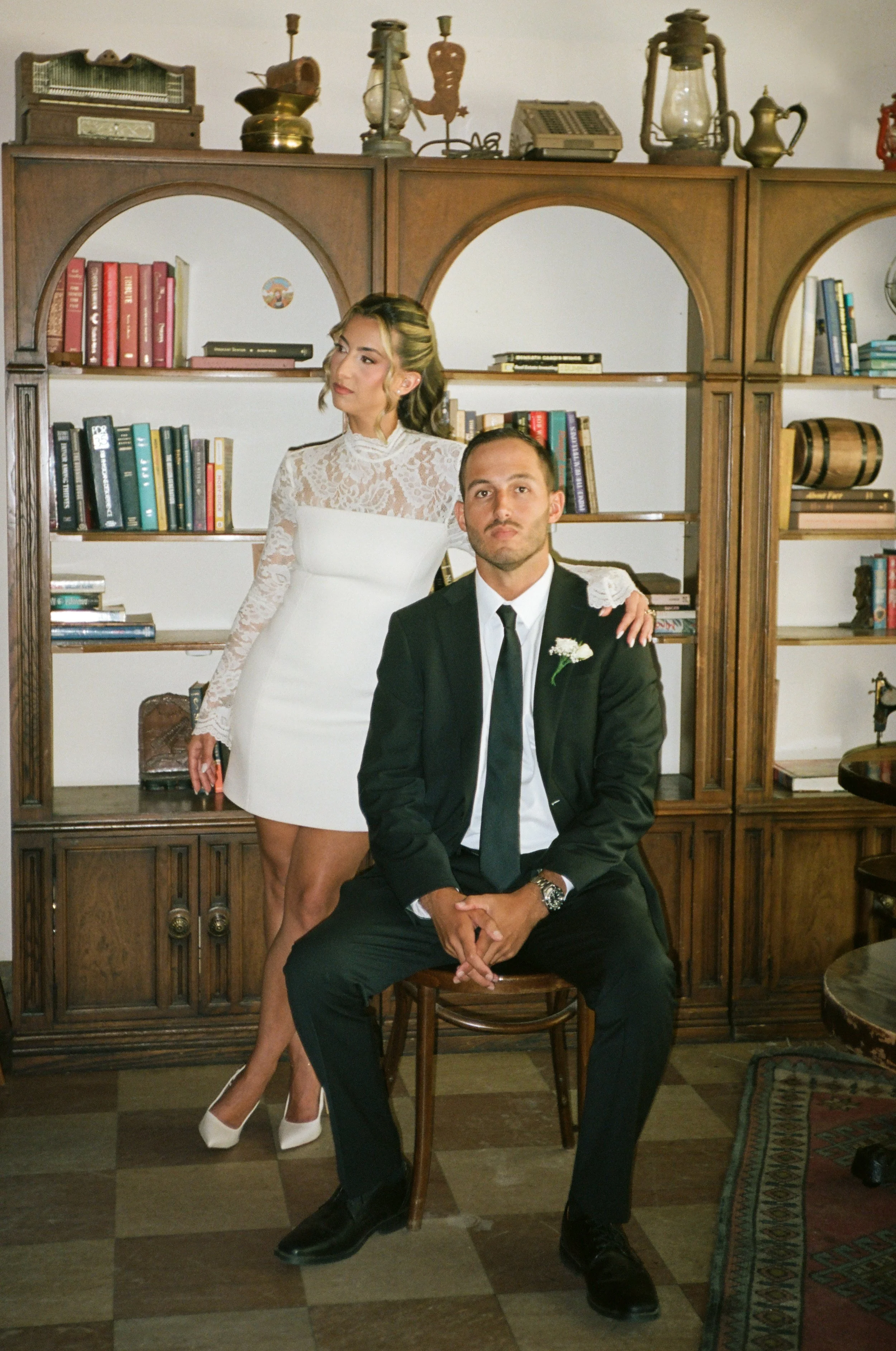 A woman in a white lace dress and heels stands behind a seated man in a black suit, white shirt, and black tie in front of a bookshelf with vintage items above it.