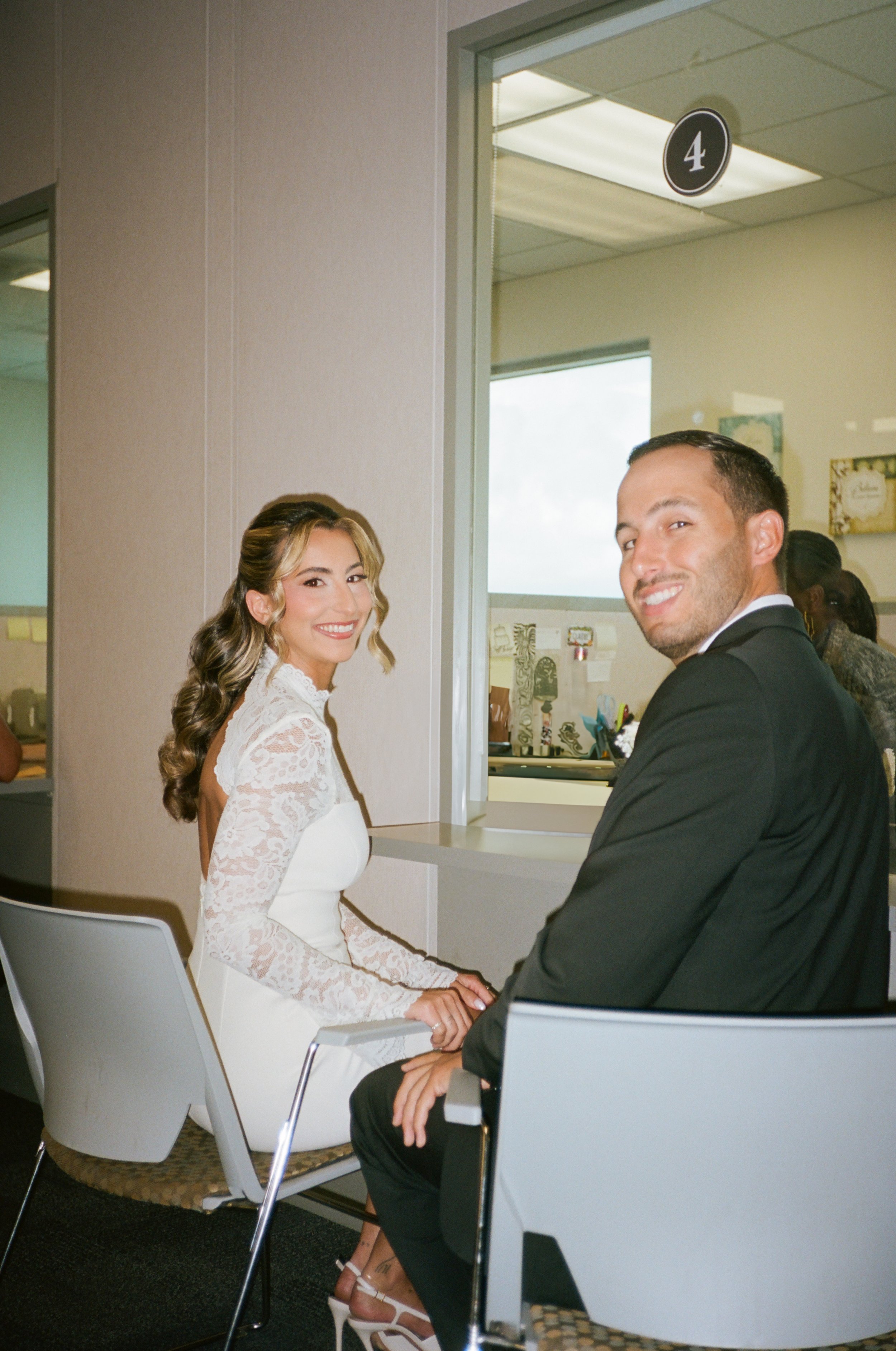 A bride and groom are holding hands while sitting in chairs in a room, likely during their wedding ceremony. The bride has blond hair, is wearing a white lace wedding dress, and white high heels. The groom has short dark hair, is dressed in a black s