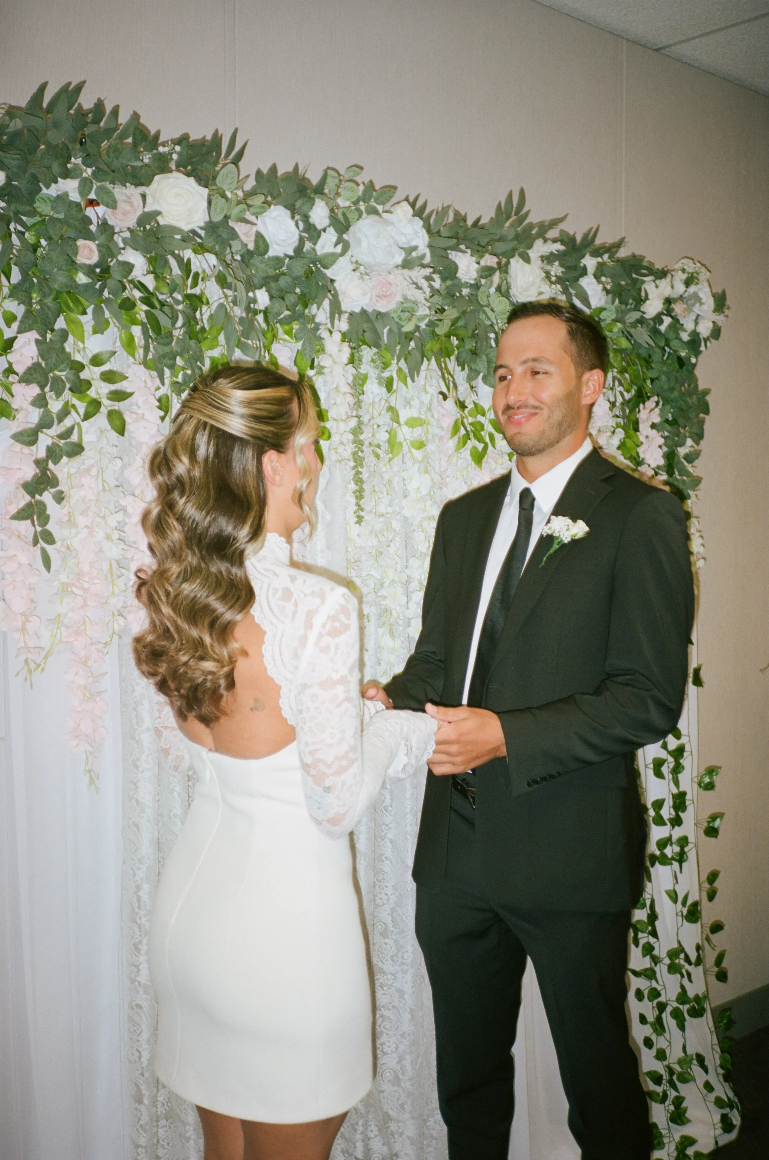 A couple stands facing each other during a wedding ceremony. The bride wears a white lace dress and has long, wavy hair, while the groom wears a black suit and tie with a boutonniere. They are holding hands in front of a floral backdrop decorated wit