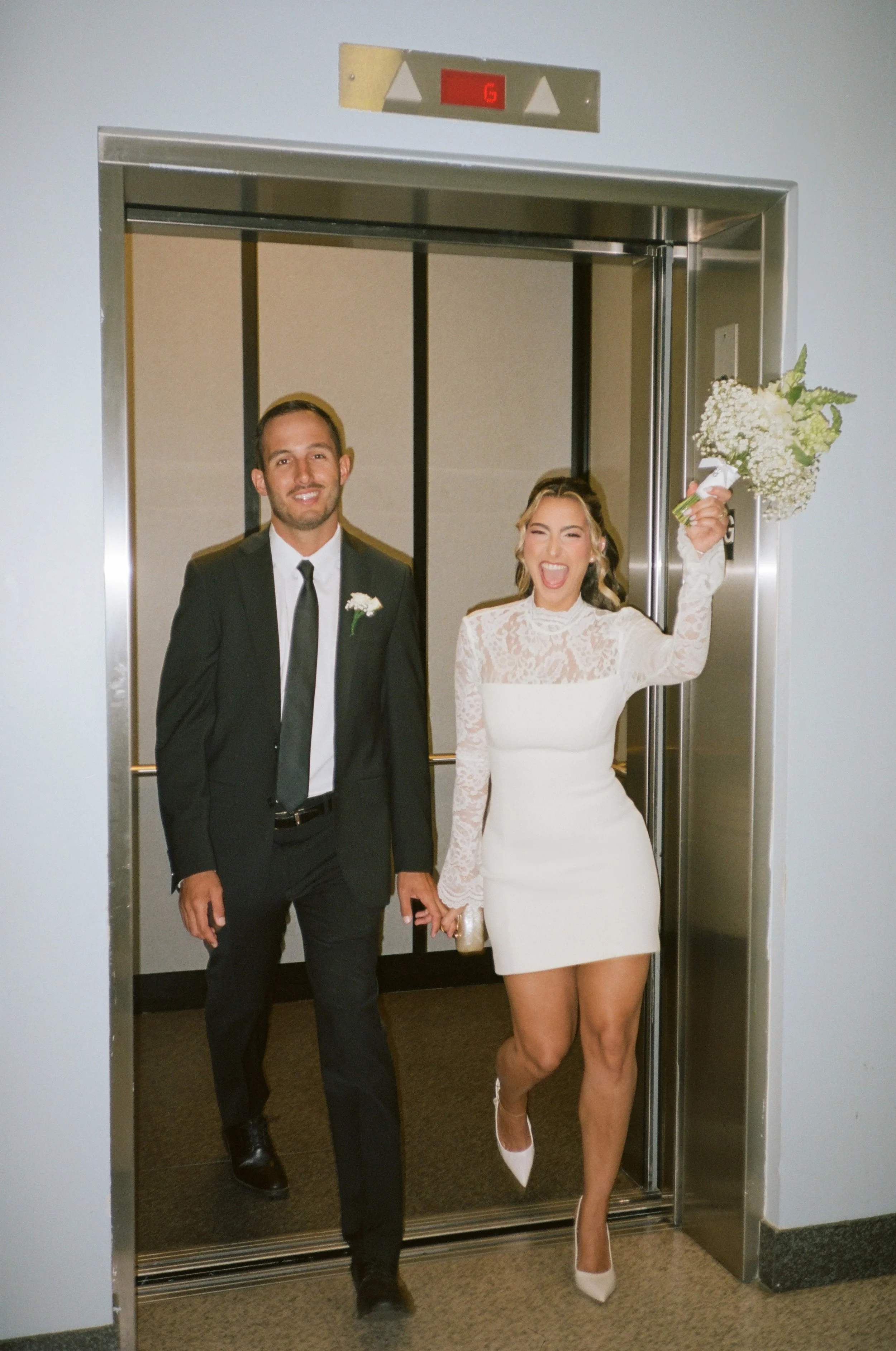 A man and woman in wedding attire holding hands and smiling as they exit an elevator.