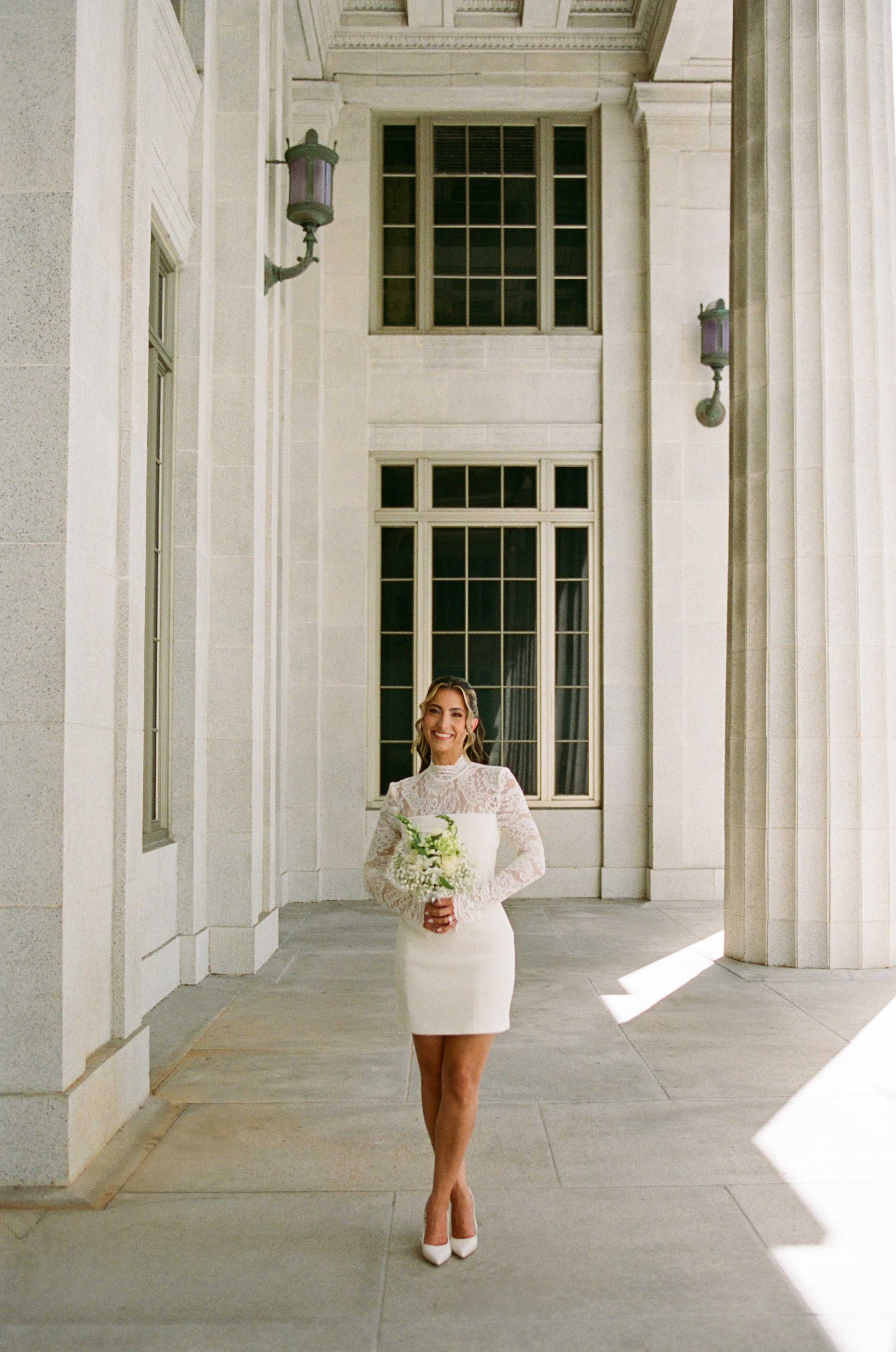 A smiling woman in a white lace dress and white heels holding a bouquet of white flowers, standing under a stone archway with tall windows and ornate lanterns.