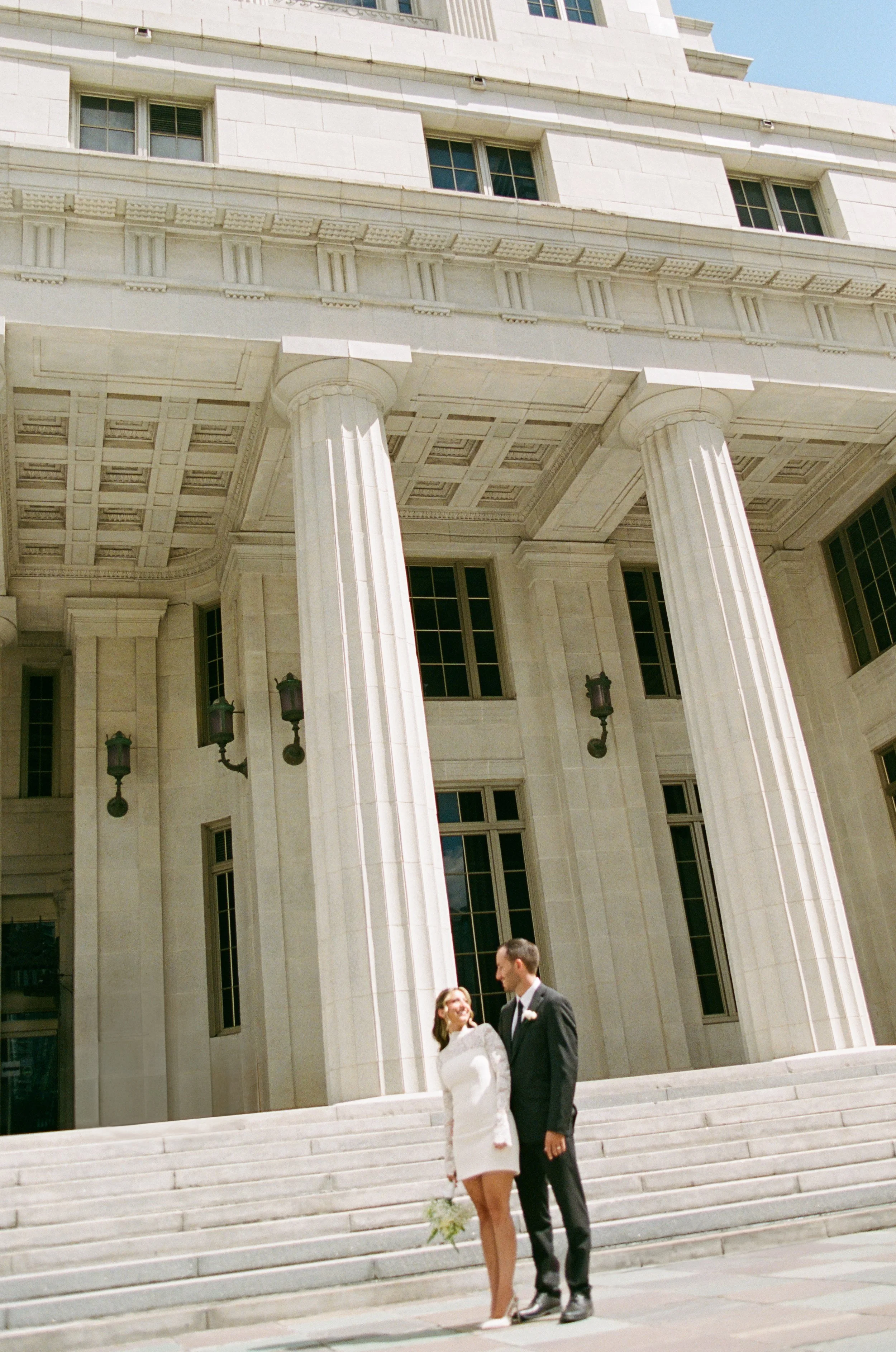 A bride and groom standing on steps outside a large classical building with columns and steps. The bride is in a white dress holding a bouquet, and the groom is in a dark suit. They are smiling and looking at each other.