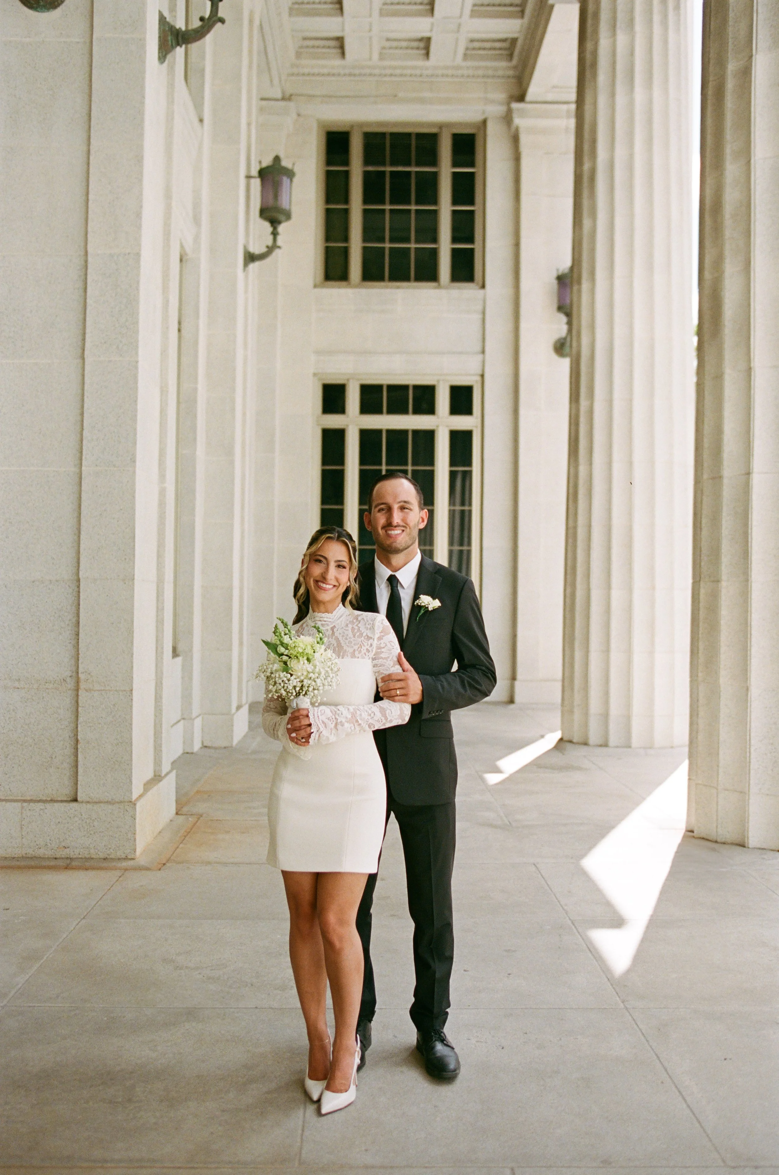 A bride and groom standing together outdoors in front of large columns and windows. The bride is wearing a white lace wedding dress and heels, holding a bouquet of white flowers, and smiling. The groom is wearing a black suit with a white shirt, blac