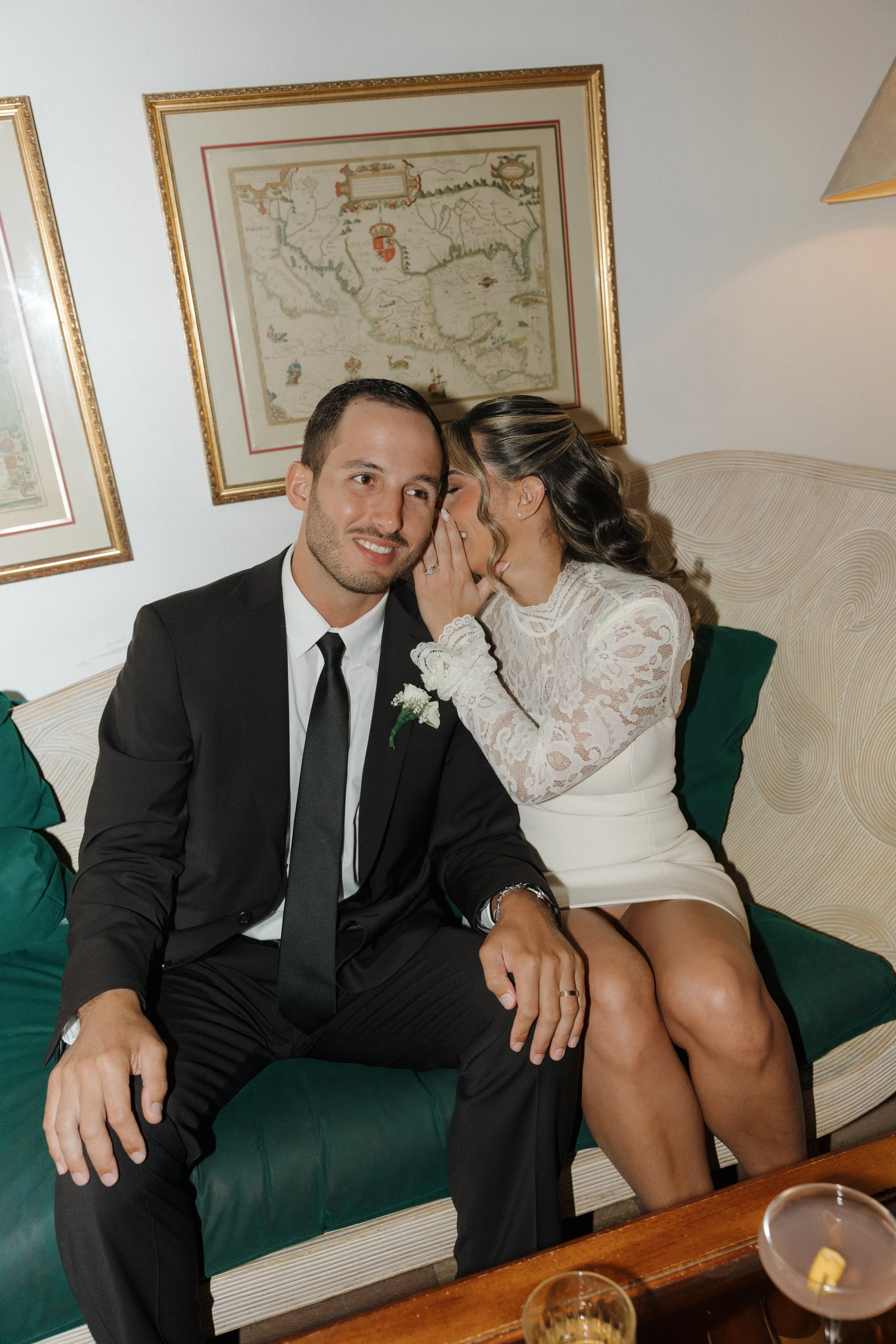 A man in a black suit and tie sitting on a green sofa, smiling, with a woman whispering into his ear, wearing a white lace dress and showing her engagement ring, in a cozy decorated room with paintings on the wall.