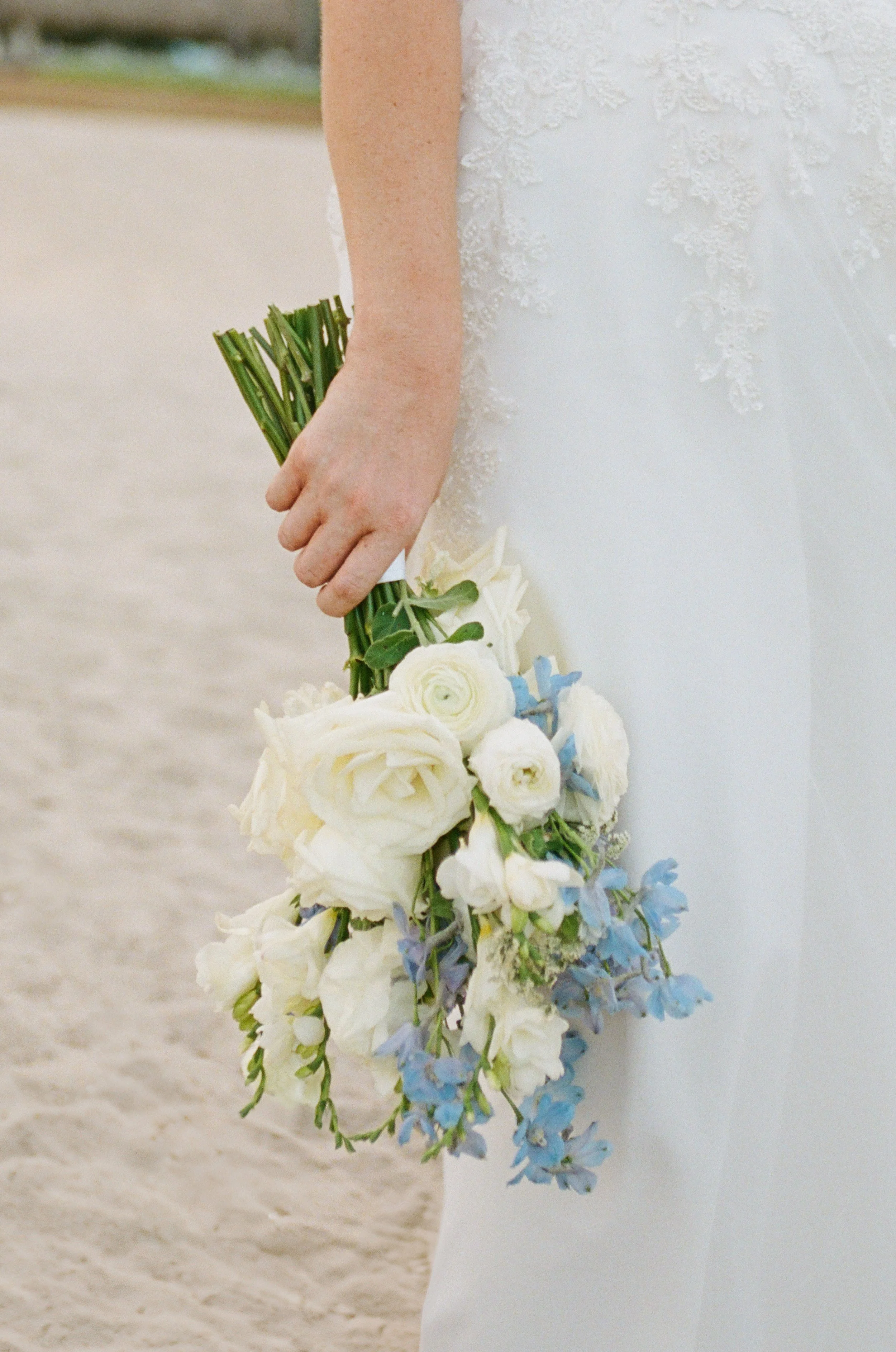 Close-up of a person in a white wedding dress holding a cascading bouquet of white and blue flowers.