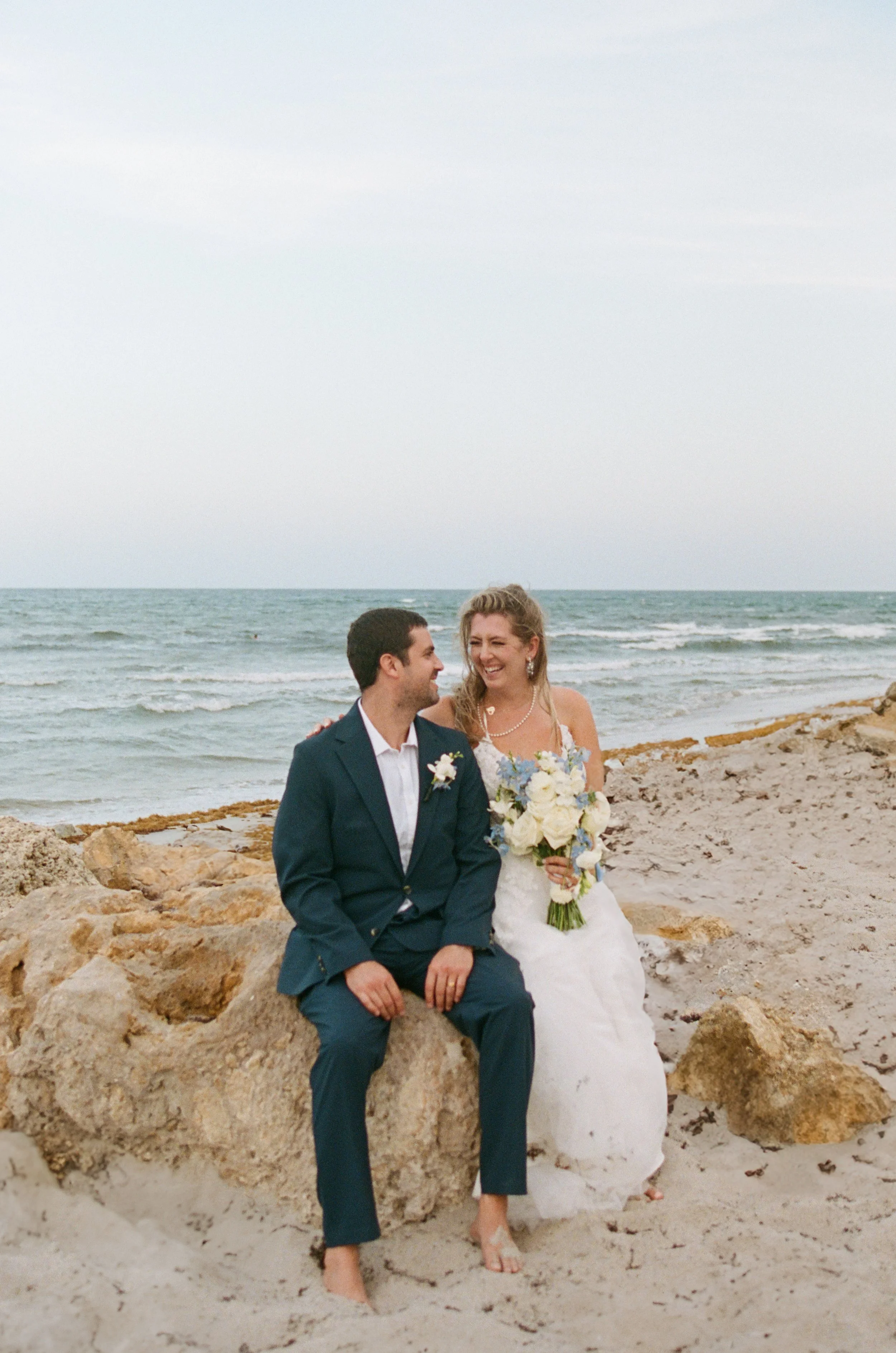 A smiling couple in wedding attire sit on rocks at the beach, with the ocean and cloudy sky in the background.