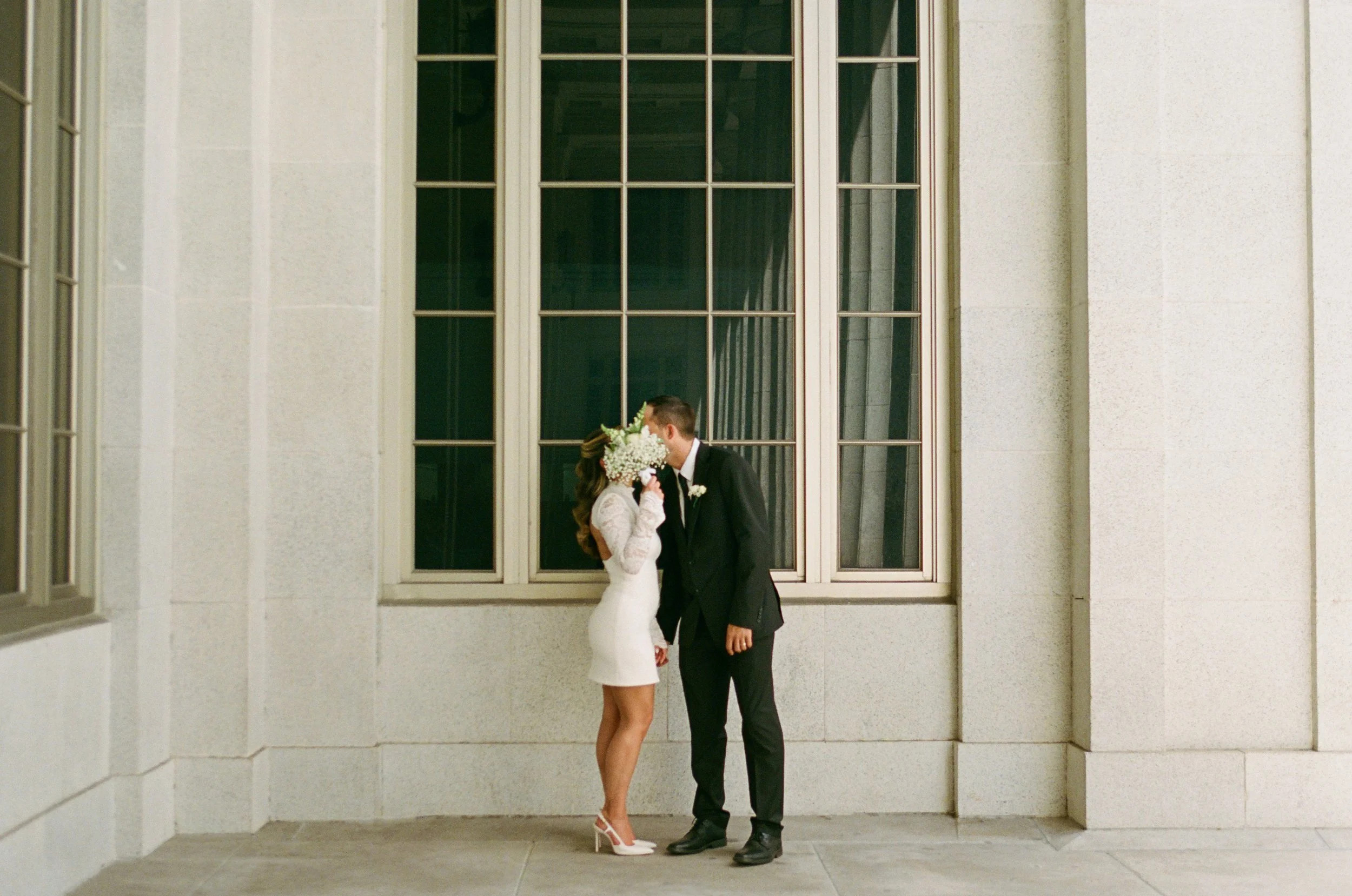 A bride and groom share a kiss in front of a large window on their wedding day. The bride wears a white dress and white heels, holding a bouquet of white flowers. The groom wears a dark suit and tie.