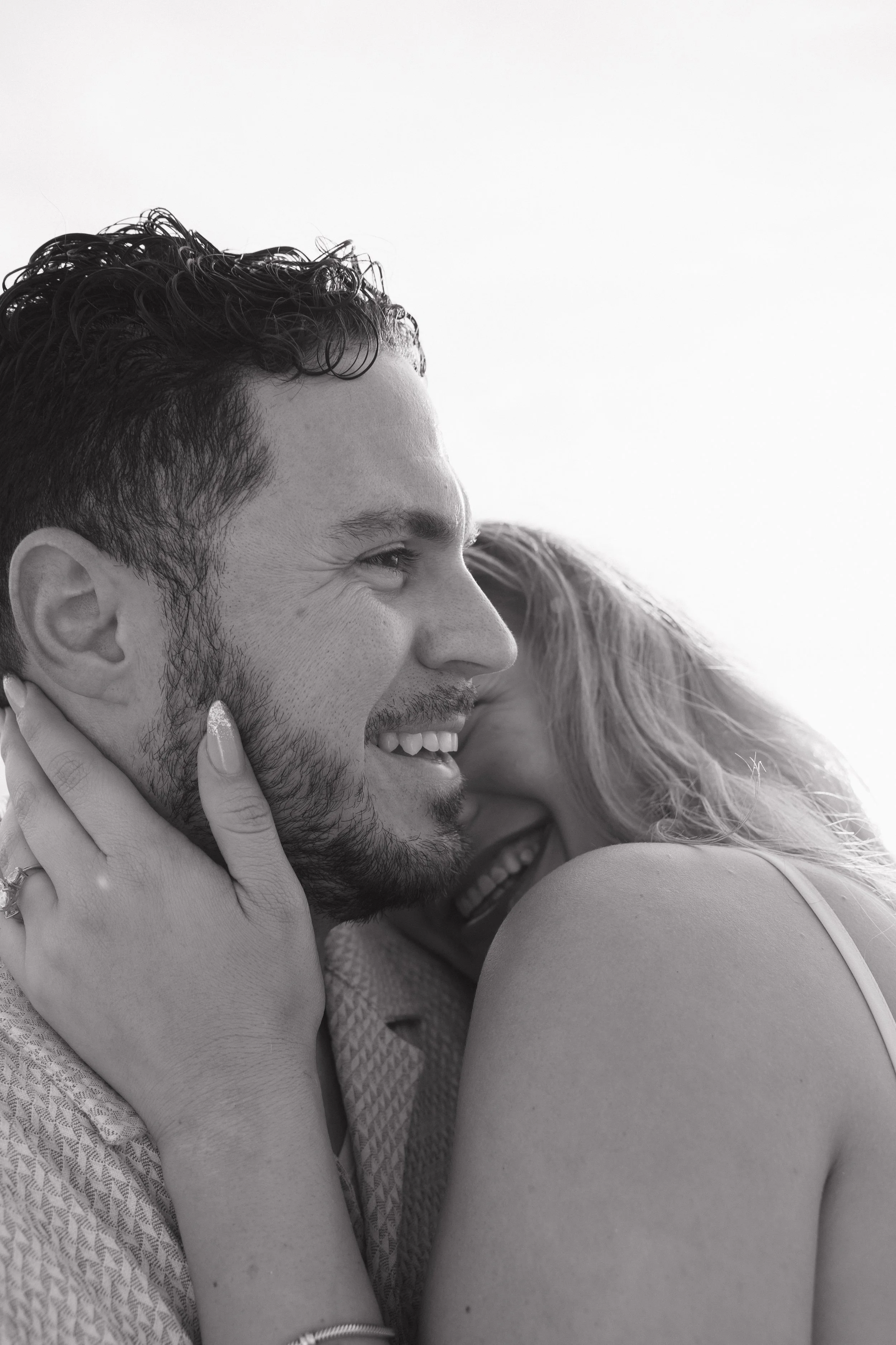 Black and white photo of a man and woman smiling and embracing. The man has curly hair and a beard, and the woman has long, wavy hair. They appear happy and close.