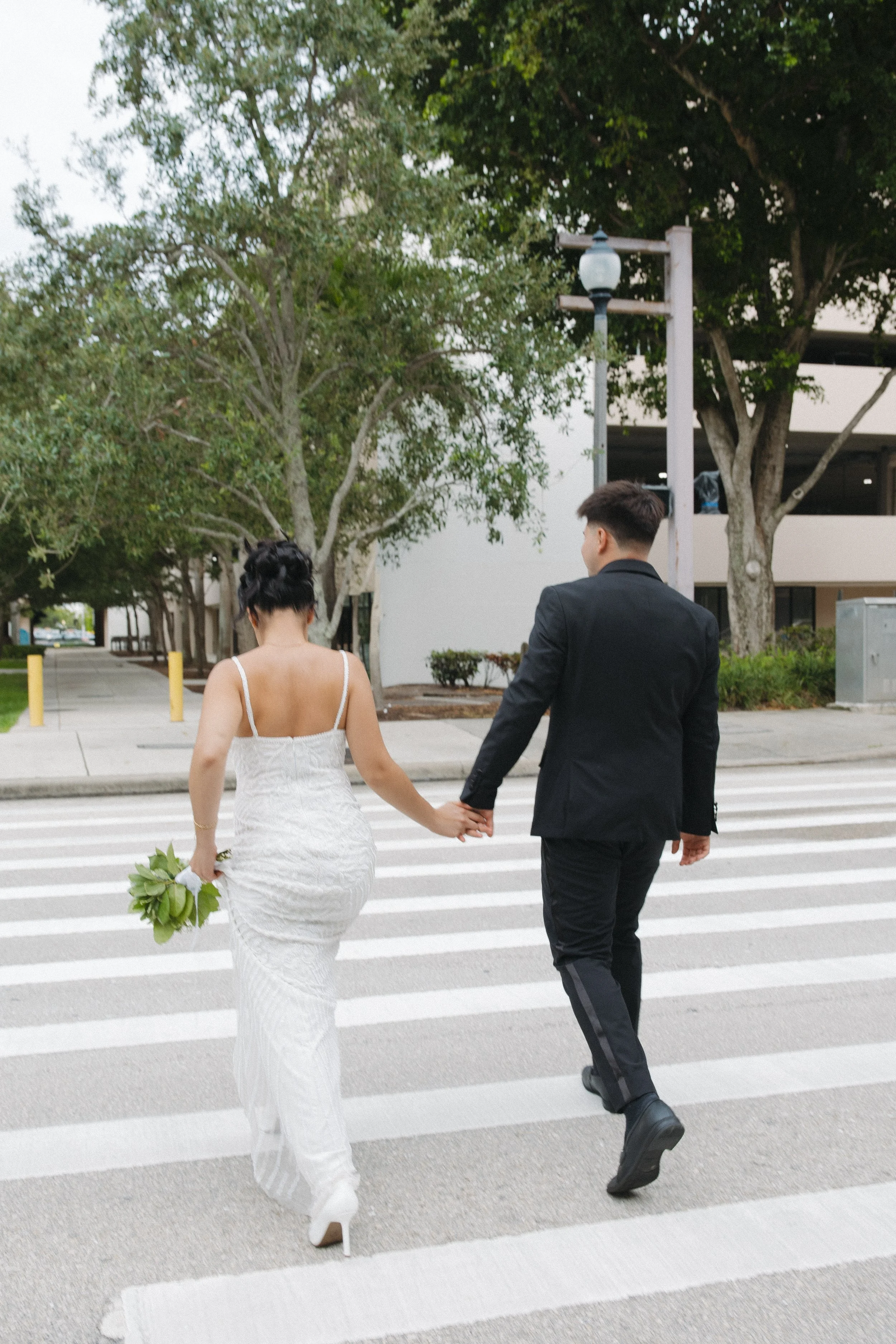 A couple walking across a crosswalk on a city street. The woman is wearing a white dress and holding a bouquet of green leaves. The man is dressed in a black suit. Trees line the street and a parking structure is visible in the background.