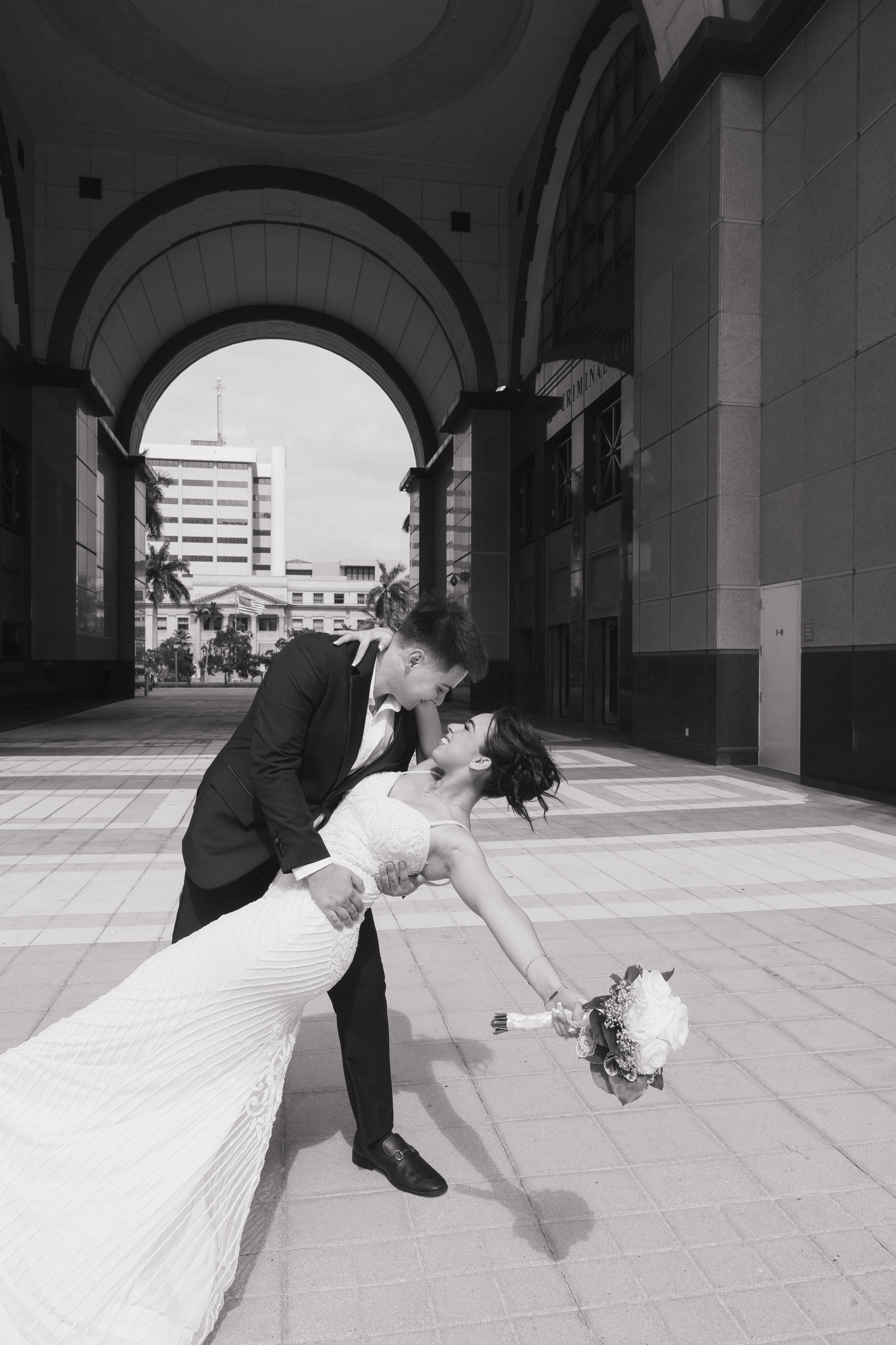 A bride and groom sharing a dance outdoors in front of a large building with arches, with the groom dipping the bride while holding her waist and she holds a bouquet of flowers, both smiling.
