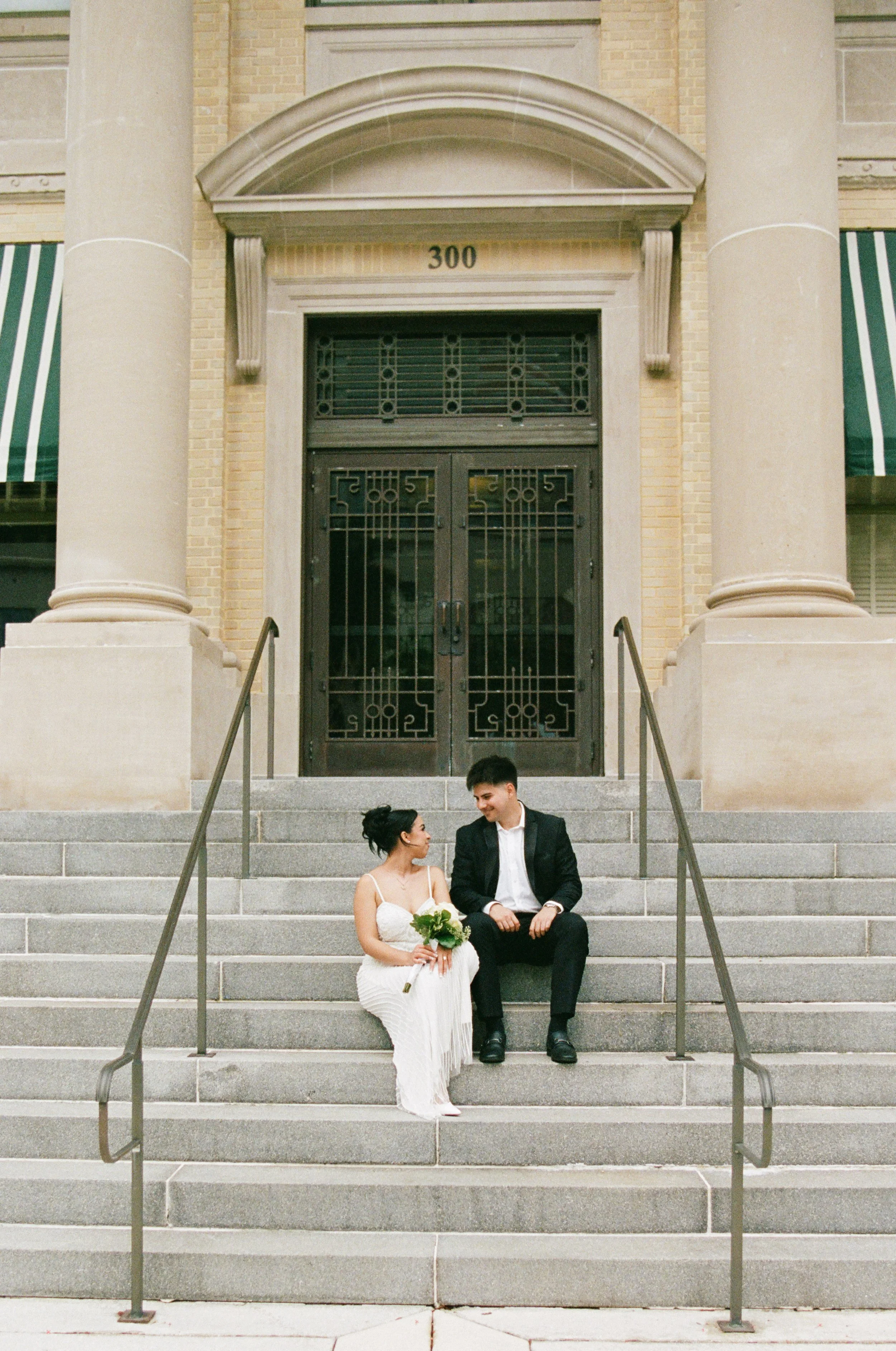 A couple sitting on the steps of a building with large columns, the woman holding a bouquet of white flowers and the man in a black suit. They are smiling at each other, dressed in formal attire, possibly for a wedding.