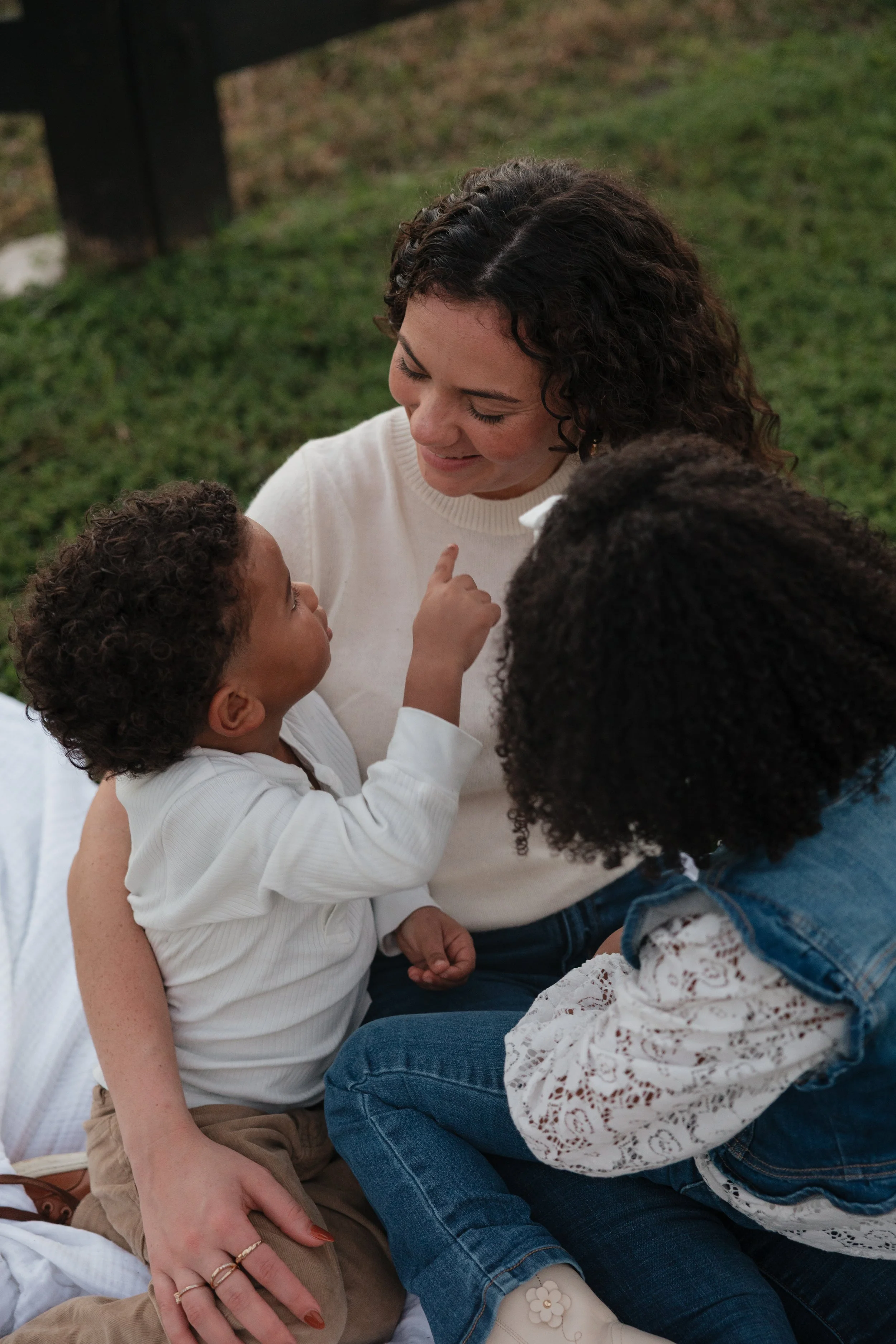 A woman with curly hair sitting on the grass, interacting affectionately with two children, one with shorter curly hair and another with long curly hair, in an outdoor setting.