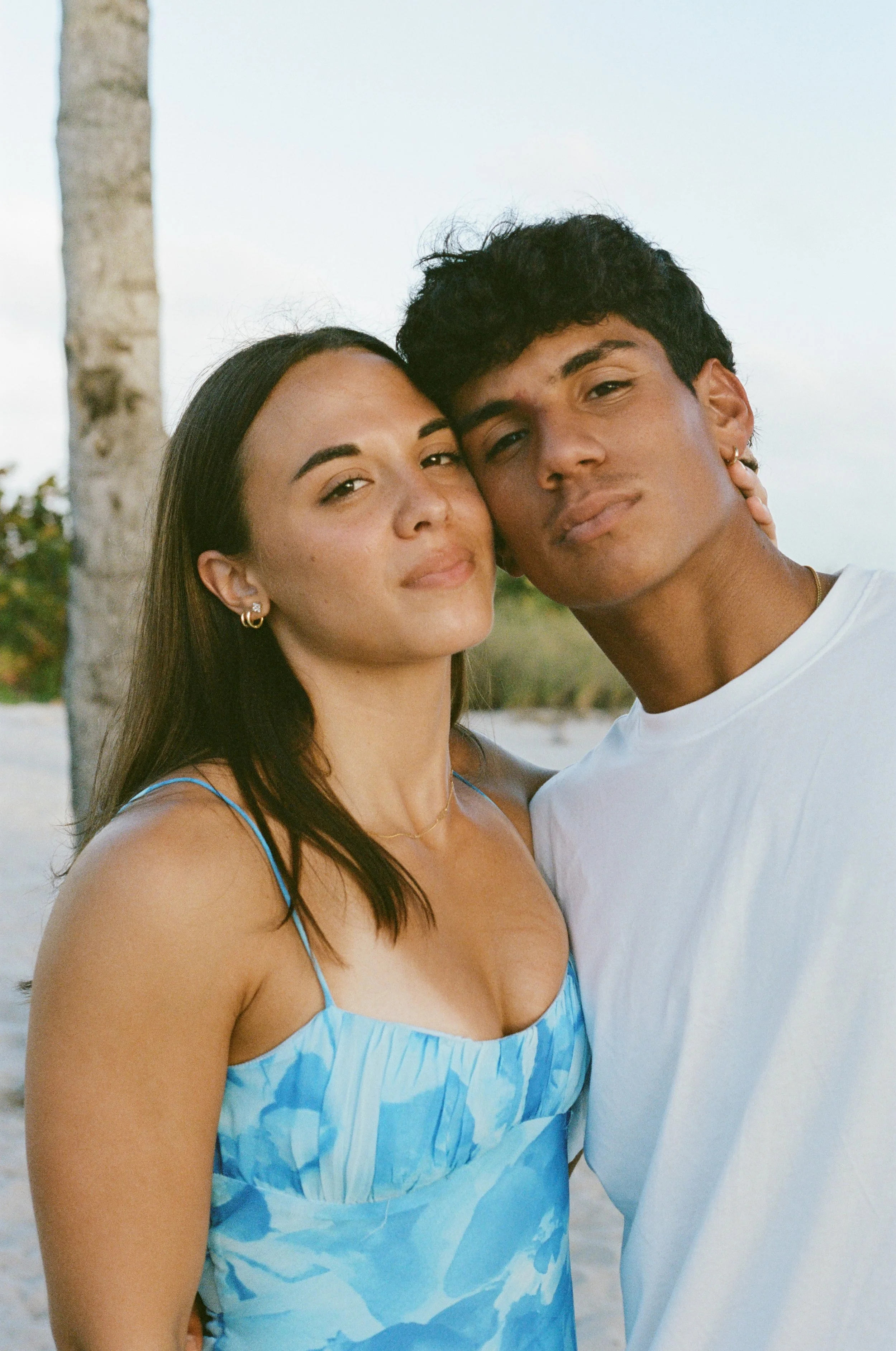 A young woman and man are standing close together outdoors during daytime, with trees and a light-colored sky in the background.