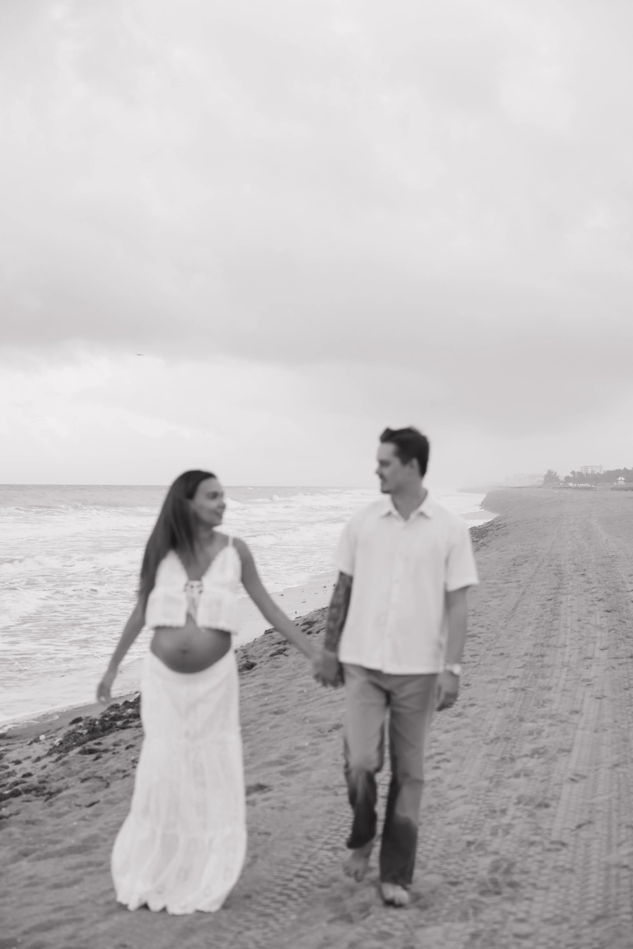 A couple walking hand in hand on a beach with the ocean and cloudy sky in the background.