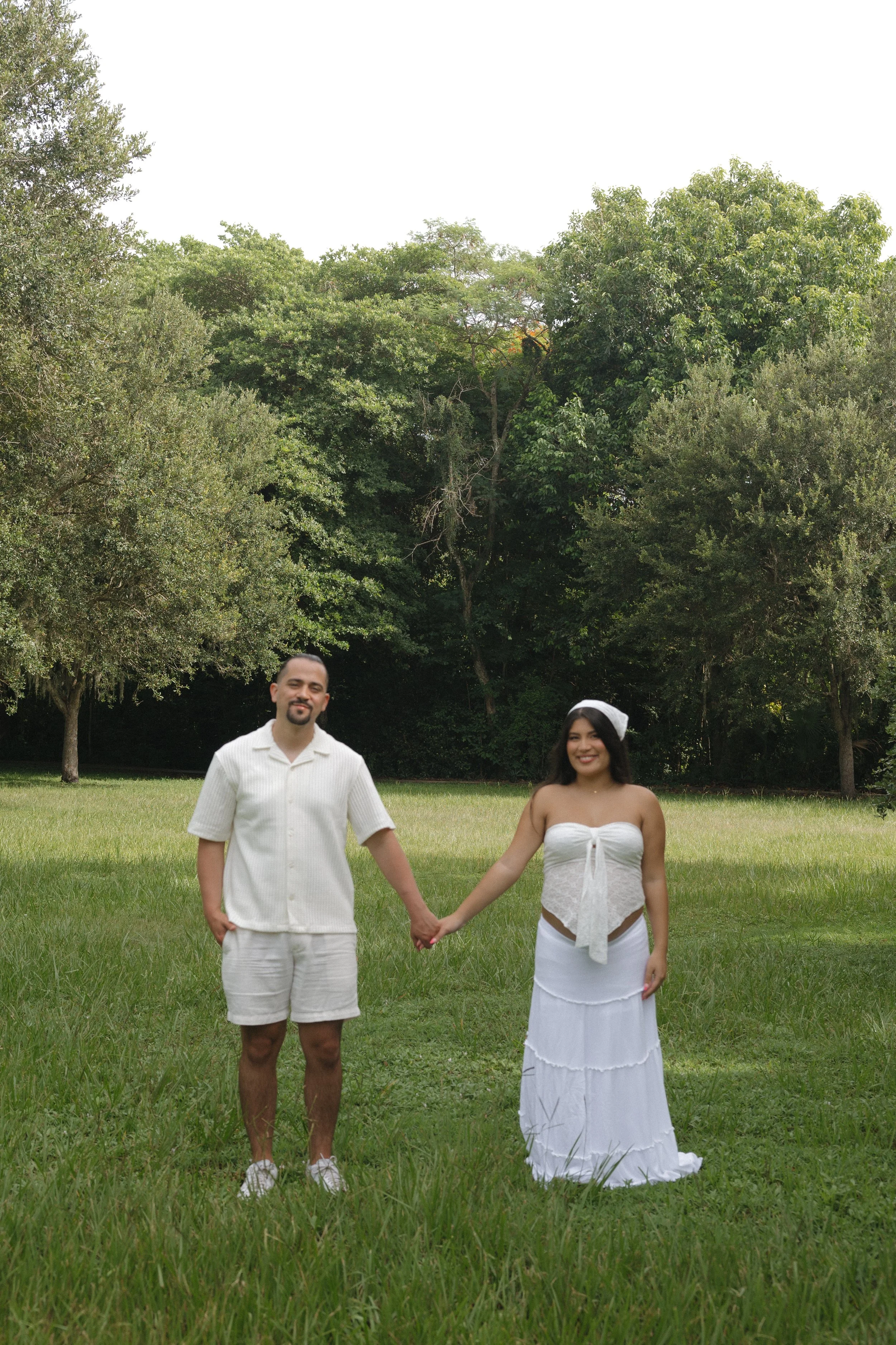 A couple holding hands, standing on a grassy field with trees in the background. The man is wearing a white short-sleeve shirt and shorts, and the woman is wearing a white strapless dress with a ribbon and a bralette underneath.