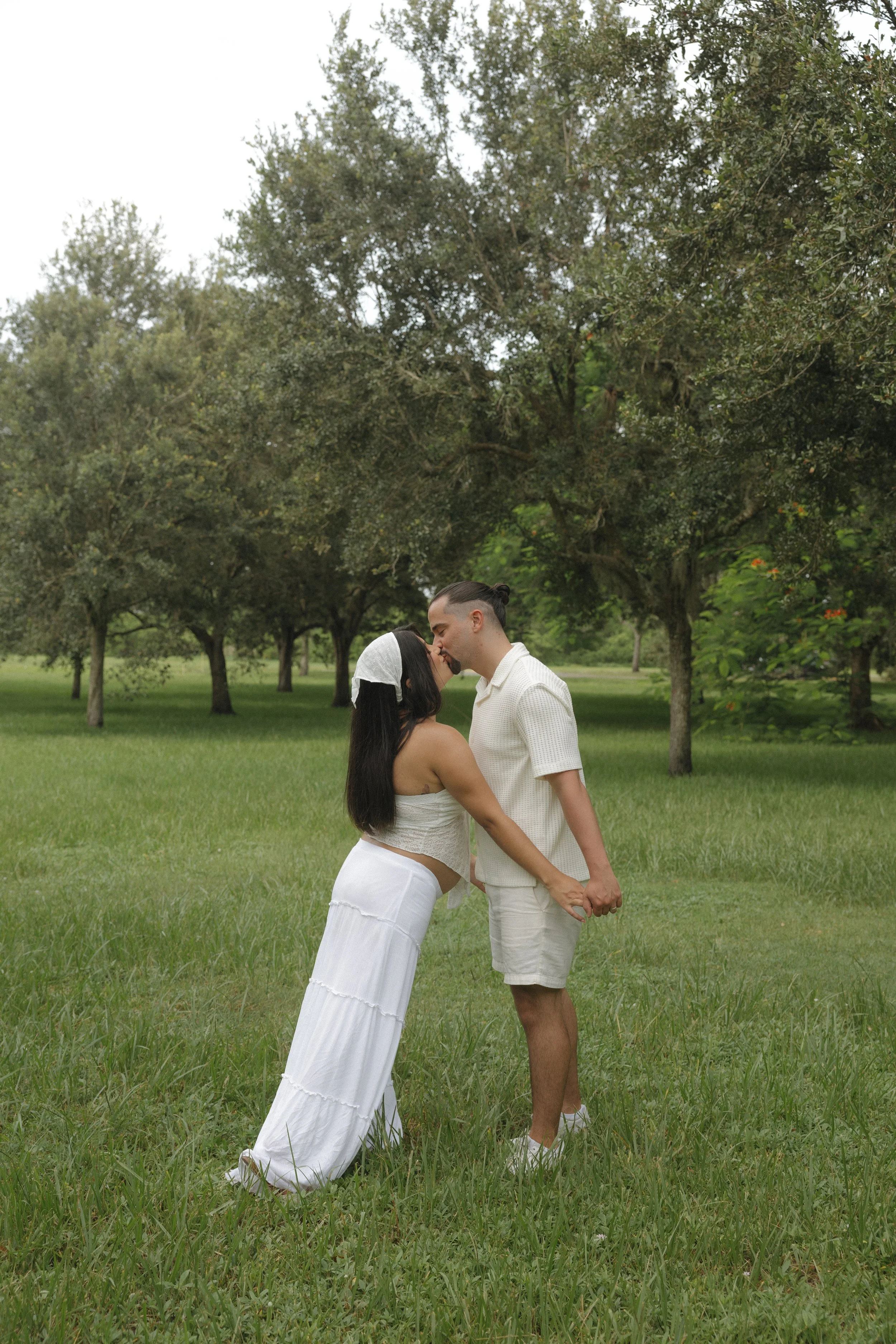 A couple holding hands and kissing in a park with green grass and trees in the background.