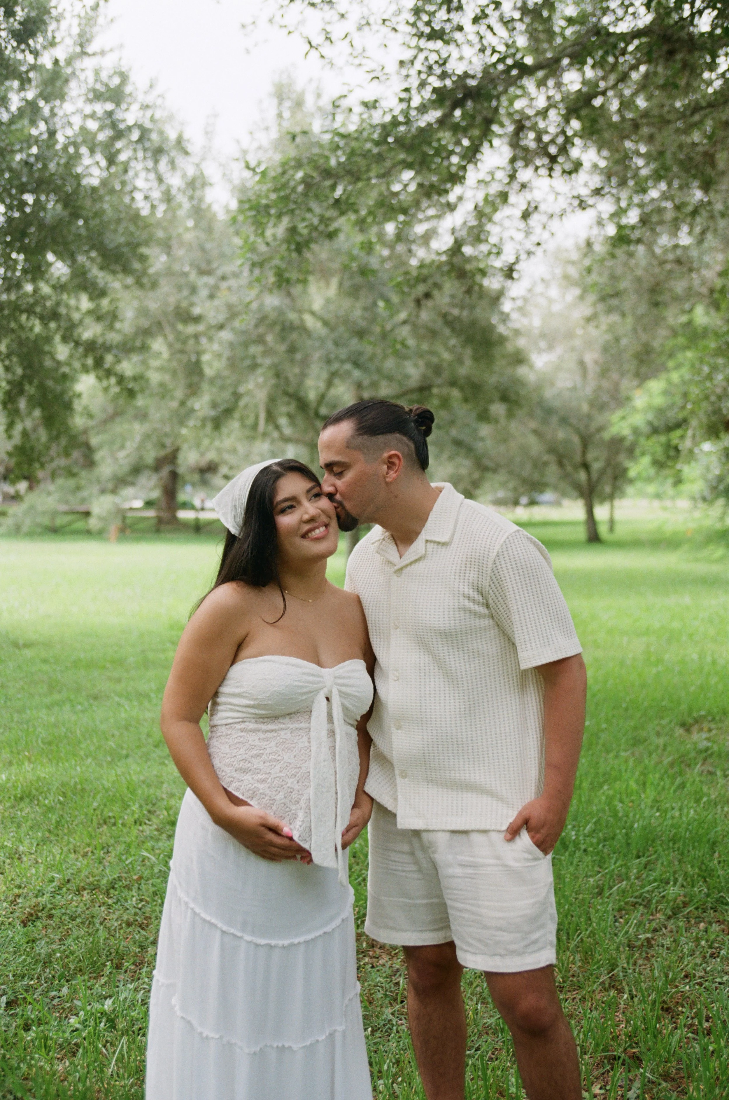 A couple stands close in a park, with the man kissing the woman's temple. The woman wears a white strapless dress and a light headscarf, smiling. The man wears a short-sleeved, light-colored shirt and shorts. Trees and green grass surround them.