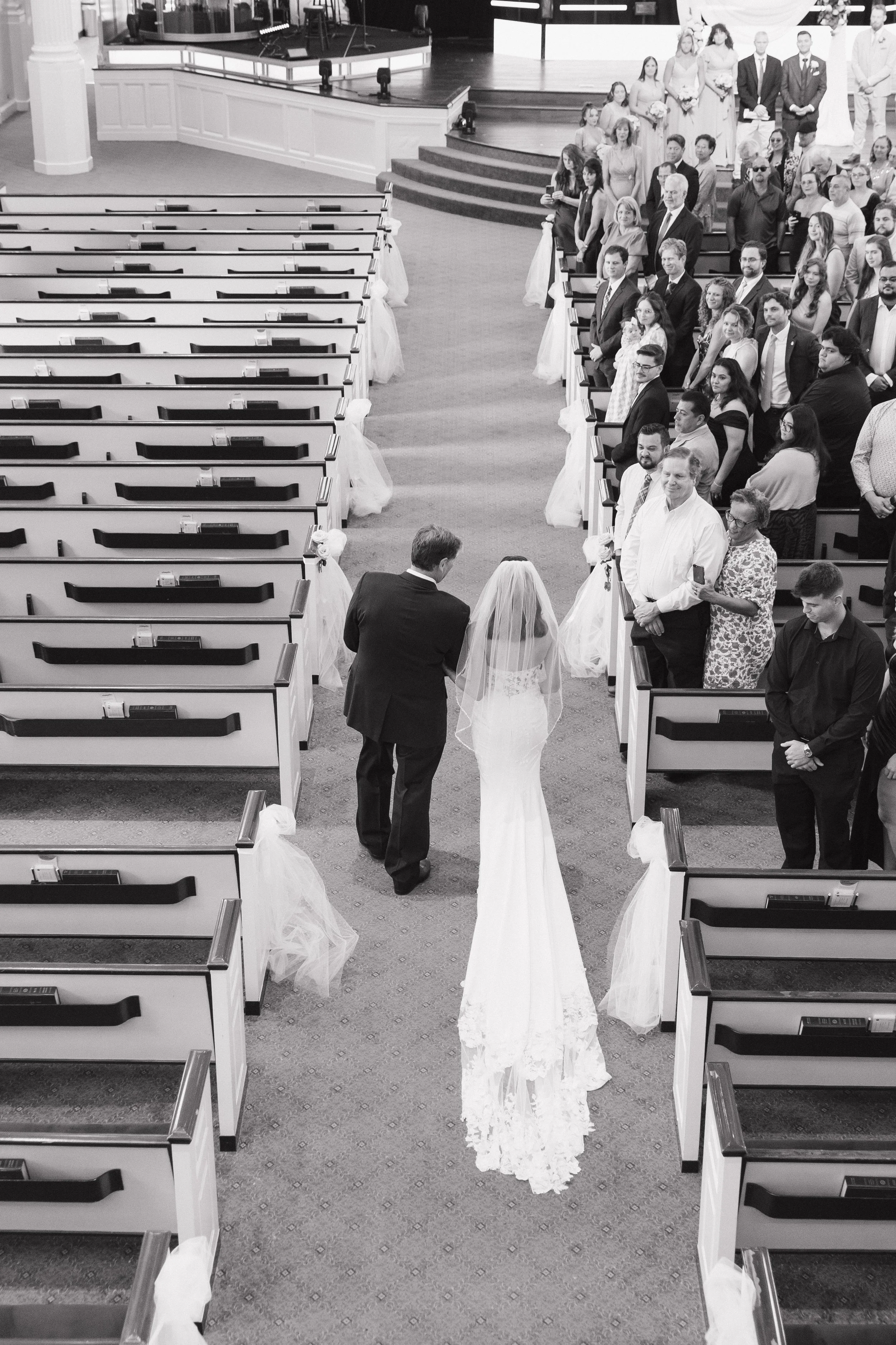 A bride walking down the aisle with a man, possibly the groom, in a wedding ceremony. Guests are standing on both sides of the aisle, watching her. The venue is decorated for a wedding with chairs, some covered with white fabric, and a stage in the b