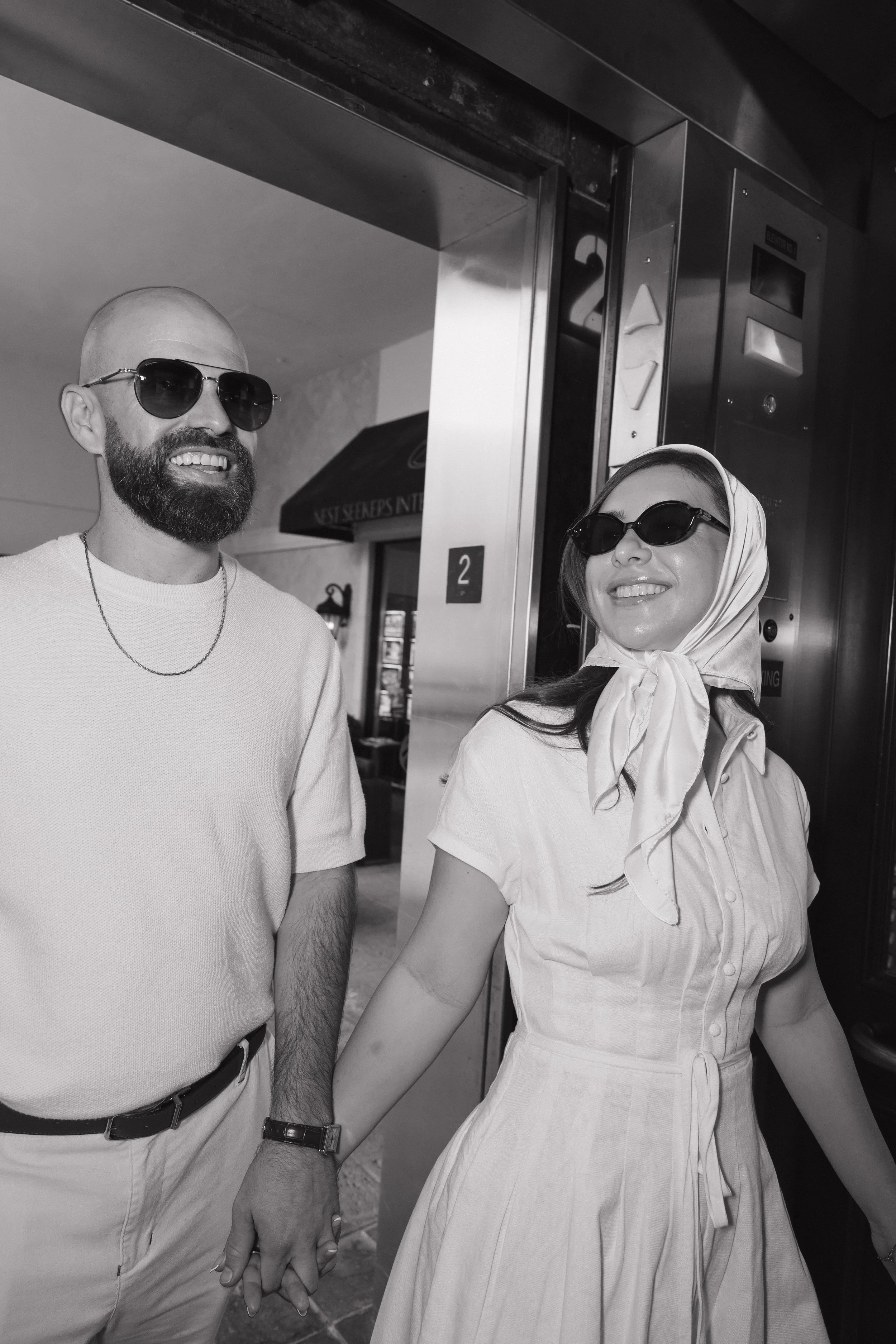 A man and a woman holding hands inside an elevator. The man is wearing sunglasses, a light-colored shirt, and a watch, with a beard and a shaved head. The woman is wearing sunglasses, a light dress, and a scarf in her hair. Both are smiling.