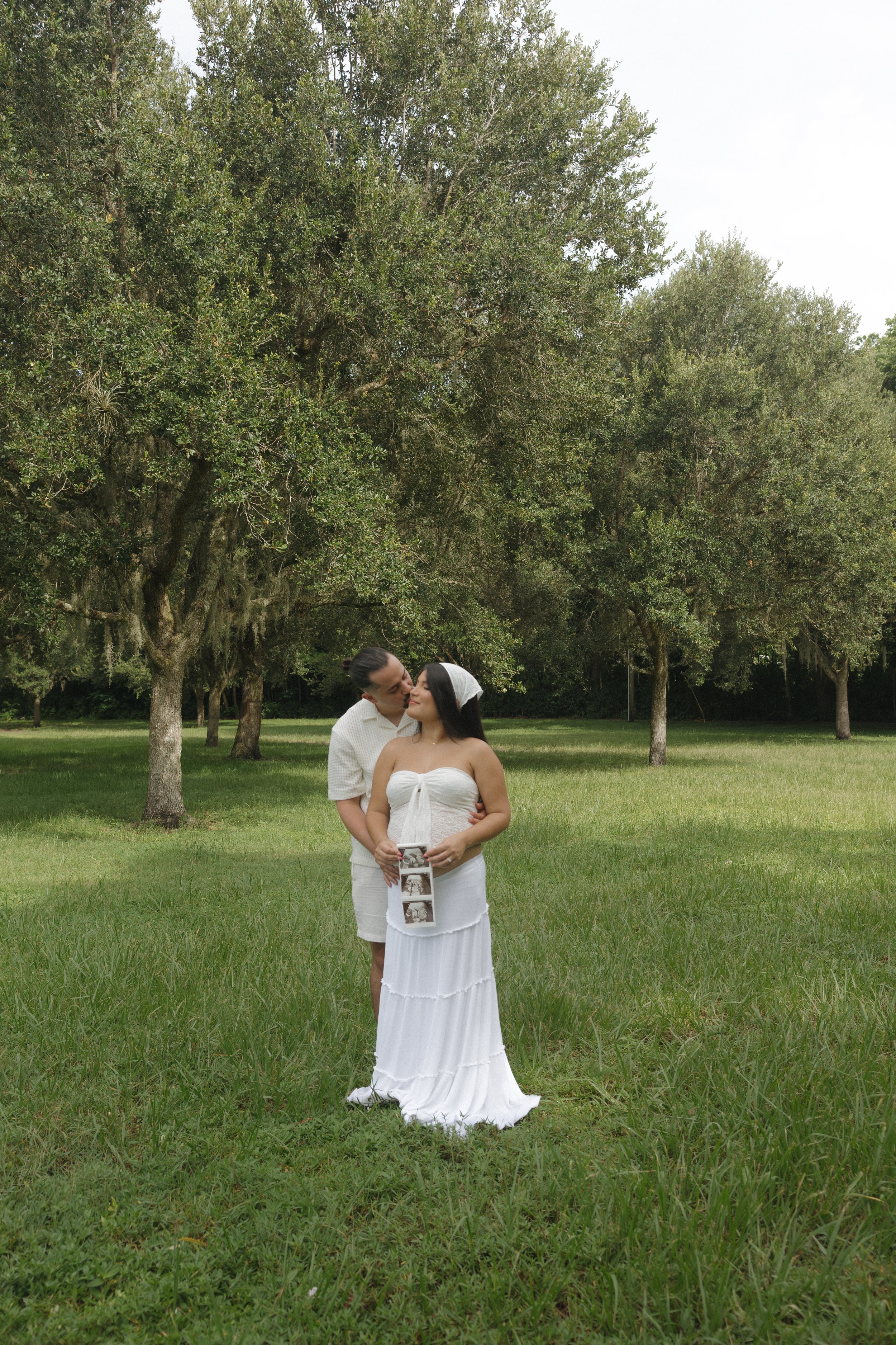 A couple standing on a grassy field holding an ultrasound photo, with the woman in a white strapless dress and the man in a light-colored shirt, surrounded by trees.