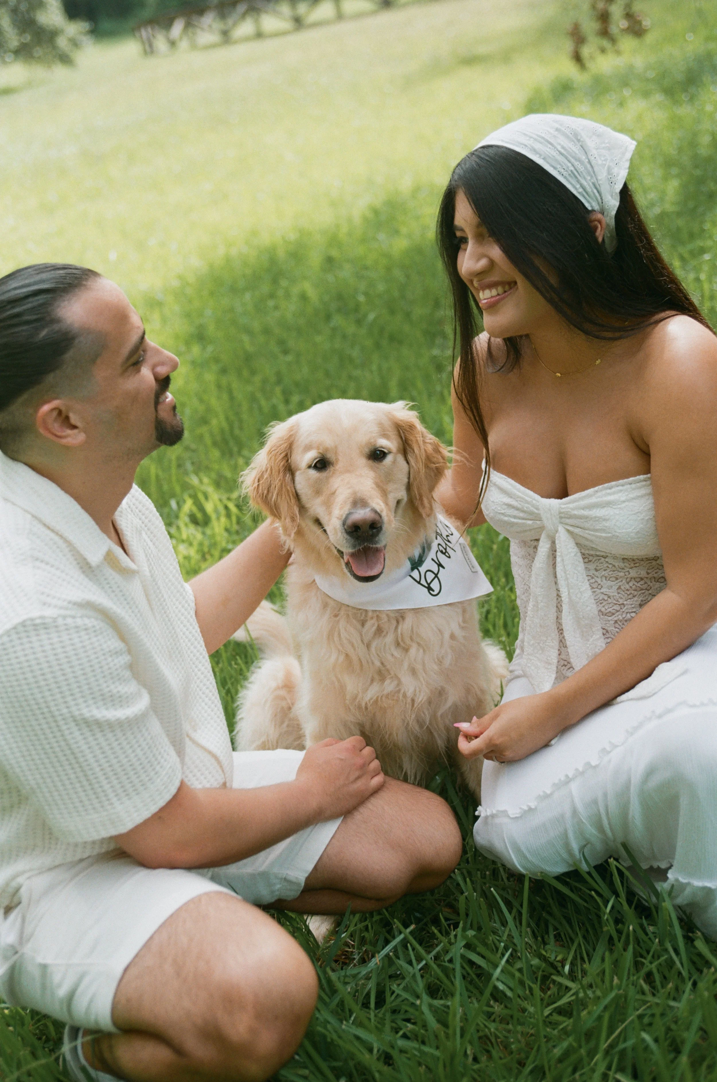 A man and woman sitting in grass outdoors, smiling at each other with a golden retriever dog between them, wearing a bandana.