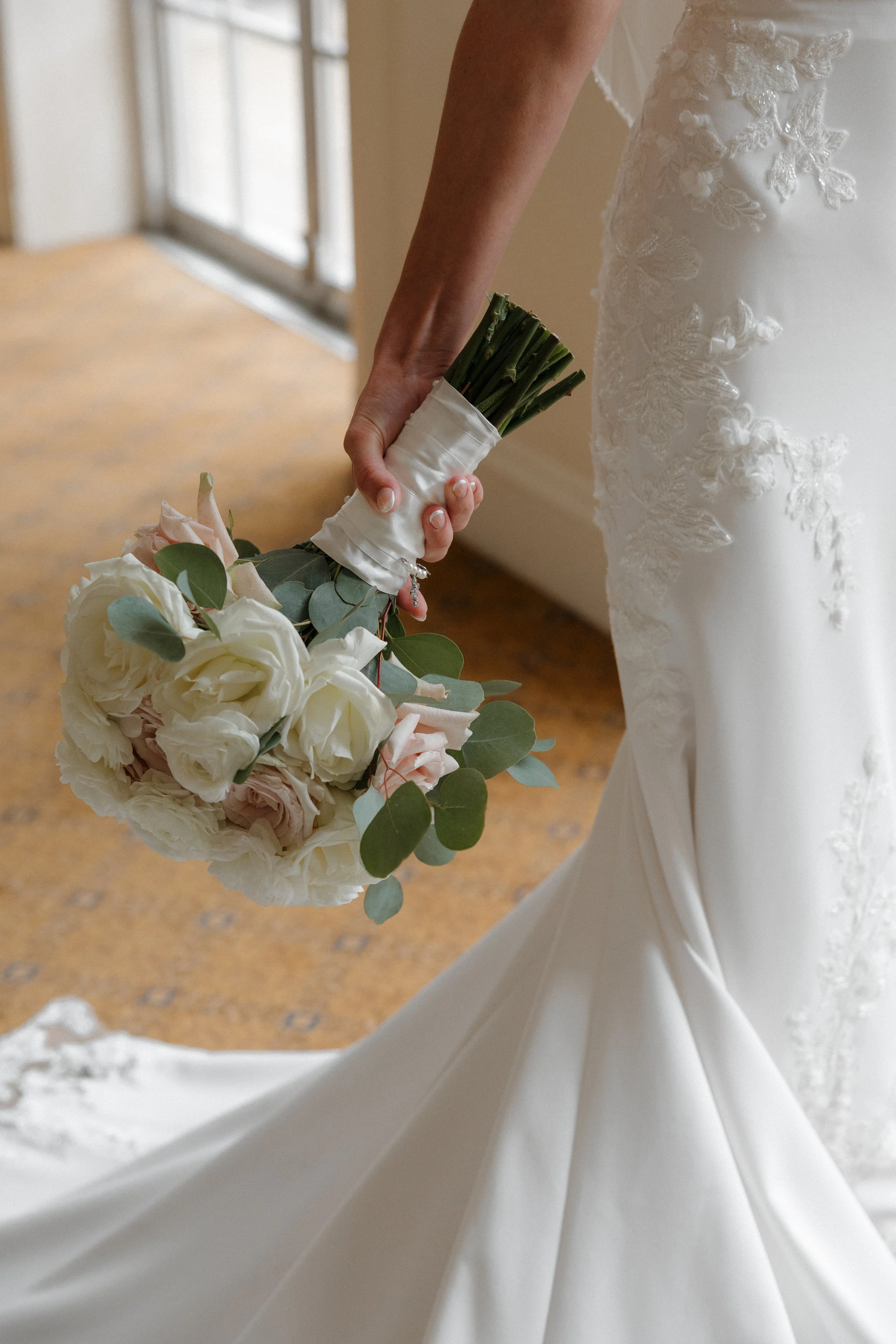 Close-up of a bride holding a bouquet of white and blush roses with eucalyptus, wearing a white wedding dress with lace embroidery, inside a room with wooden flooring and large windows.