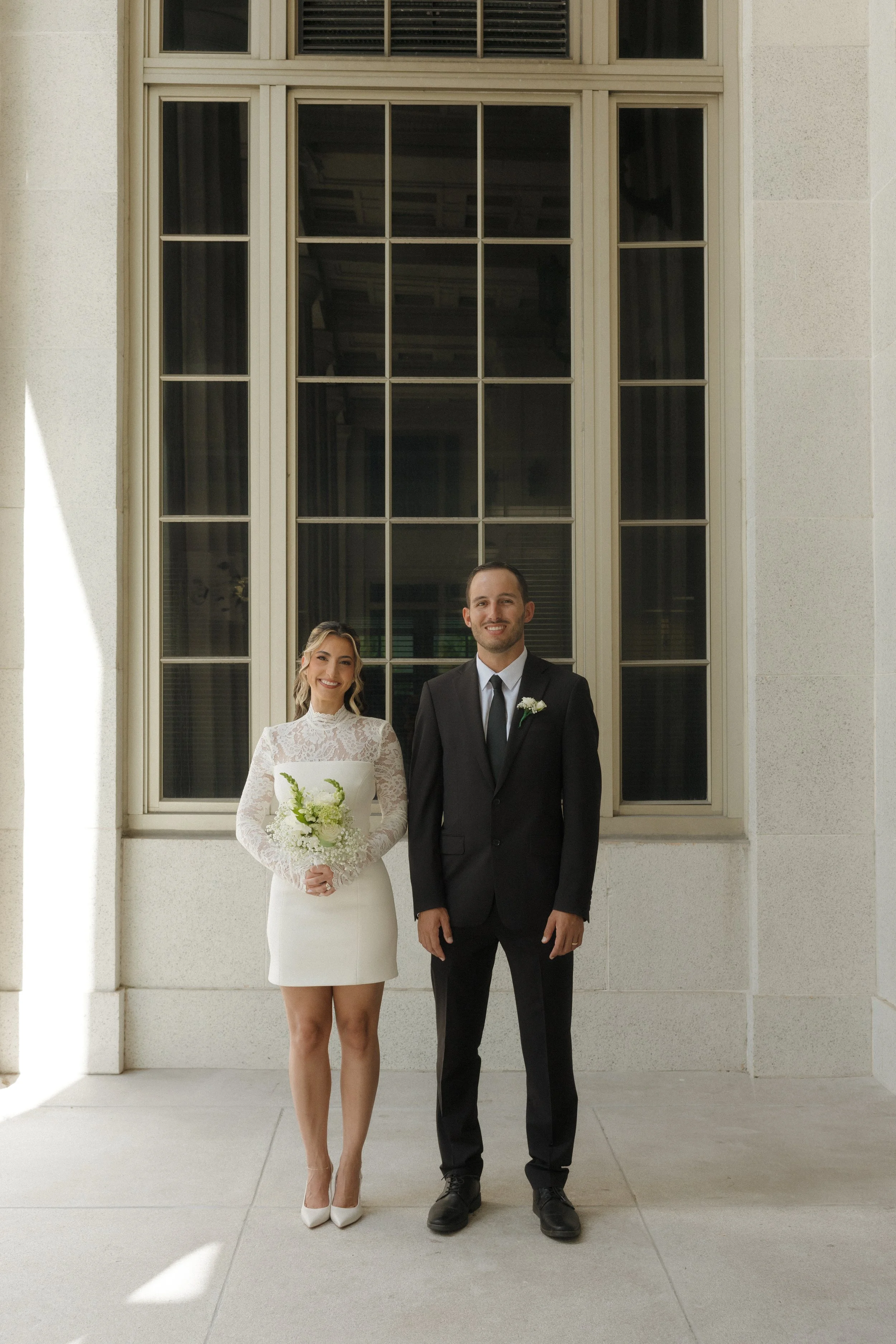 A smiling bride and groom standing side by side outside in front of a cream-colored building with large windows. The bride is holding a bouquet of white flowers and wears a white lace dress with long sleeves and high heels. The groom is in a black su