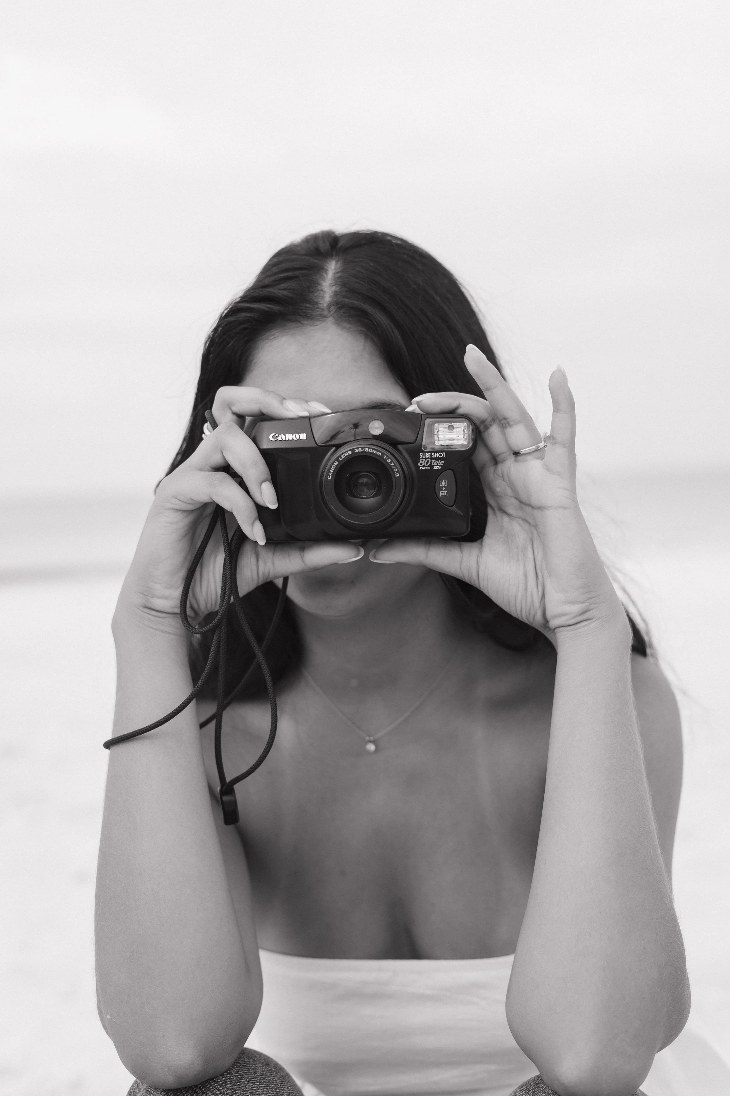 A woman with dark hair taking a photo with a Canon camera, viewed from behind, with her elbows resting on her knees, in front of a plain background.