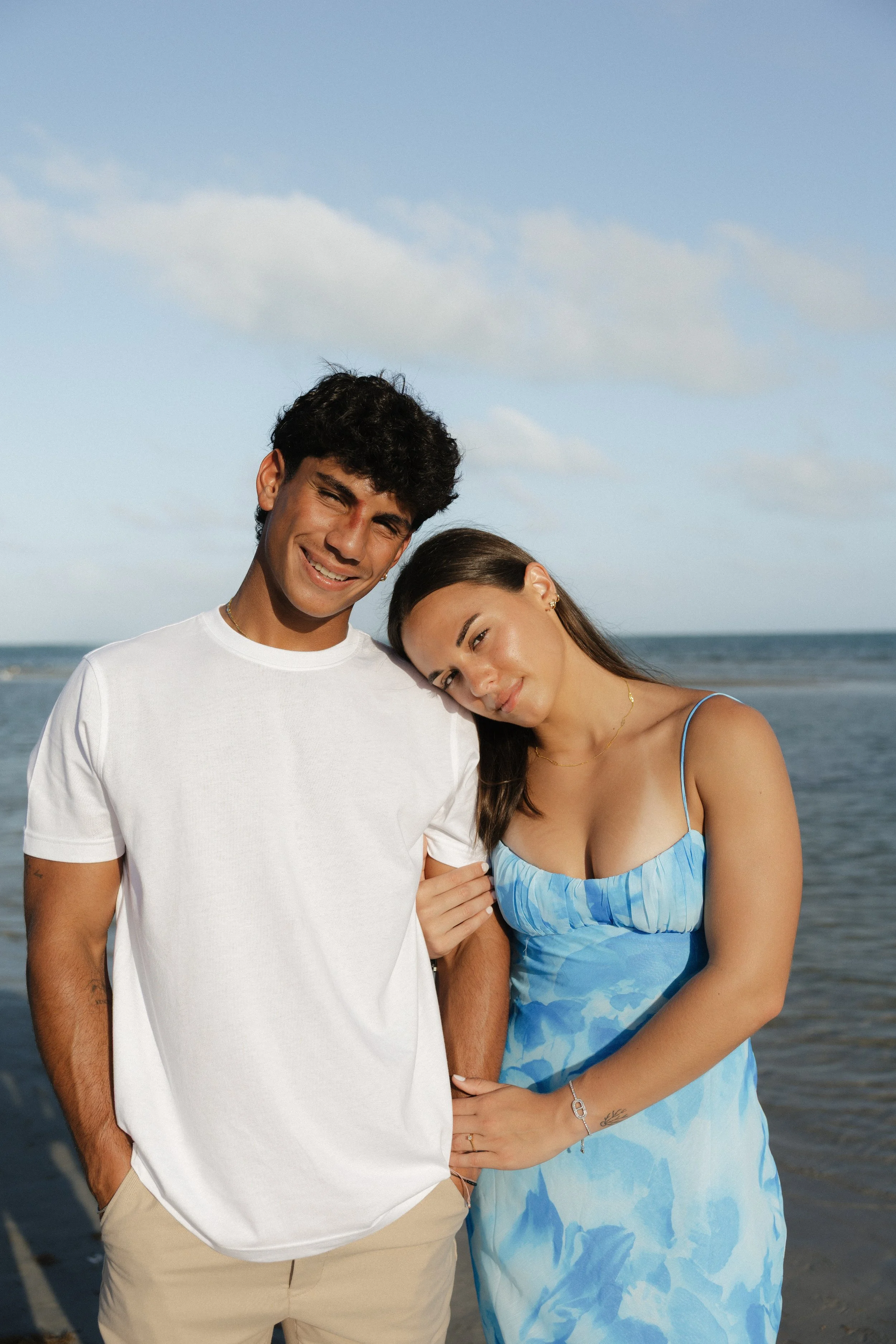 A young couple stands close together on the beach, with the ocean and a partly cloudy sky in the background. The man has dark, curly hair, is smiling, and wears a white t-shirt and beige pants. The woman, with long brown hair, leans her head on his s
