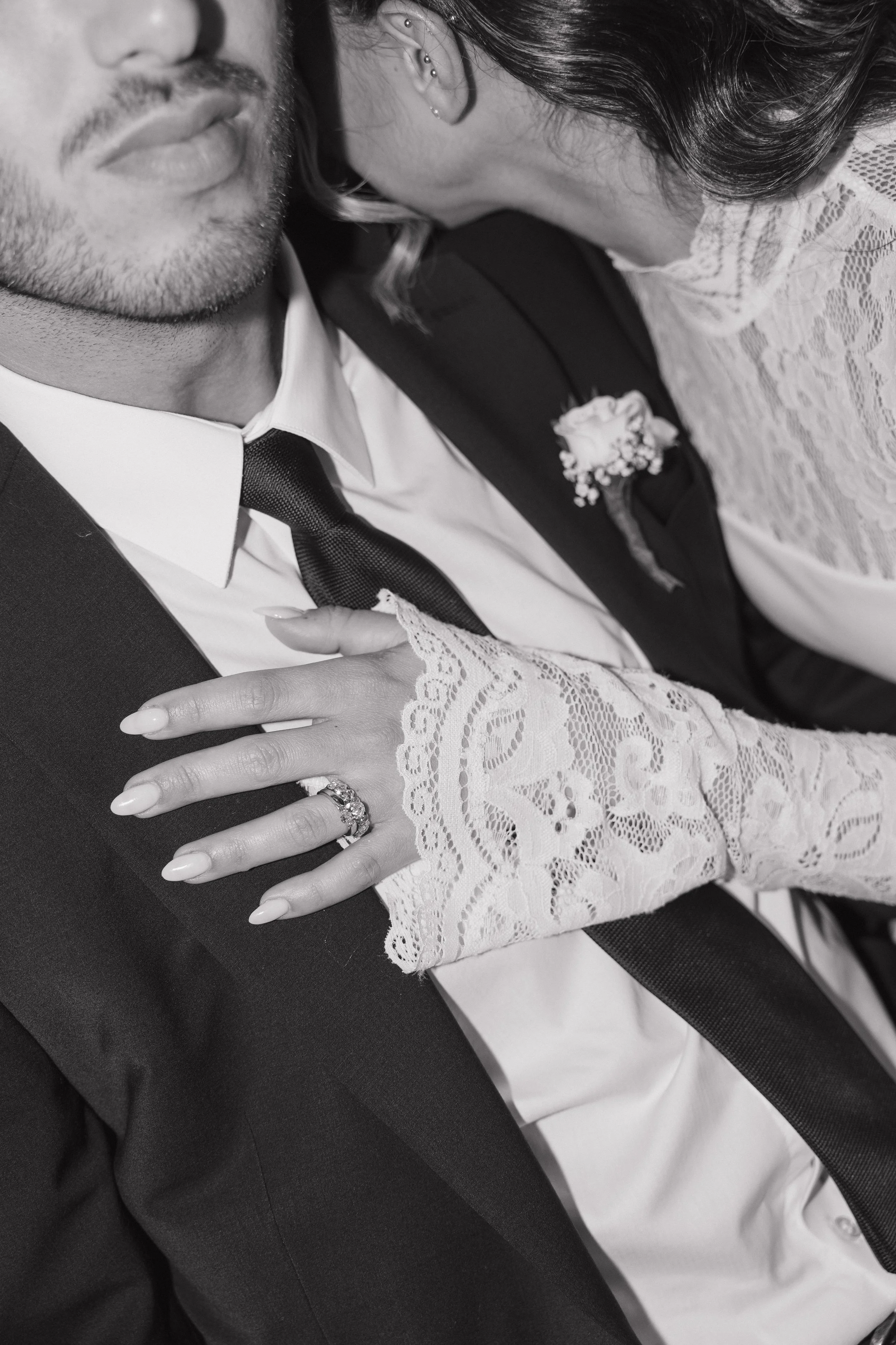 A close-up black and white photograph of a bride and groom on their wedding day. The bride is resting her head on the groom's shoulder, showing her lace dress and a wedding ring on her finger, which is placed on the groom's chest. The groom is dressed in a suit with a tie and has a boutonniere on his lapel. The bride's head is turned towards the groom, and she has multiple earrings in her ear.