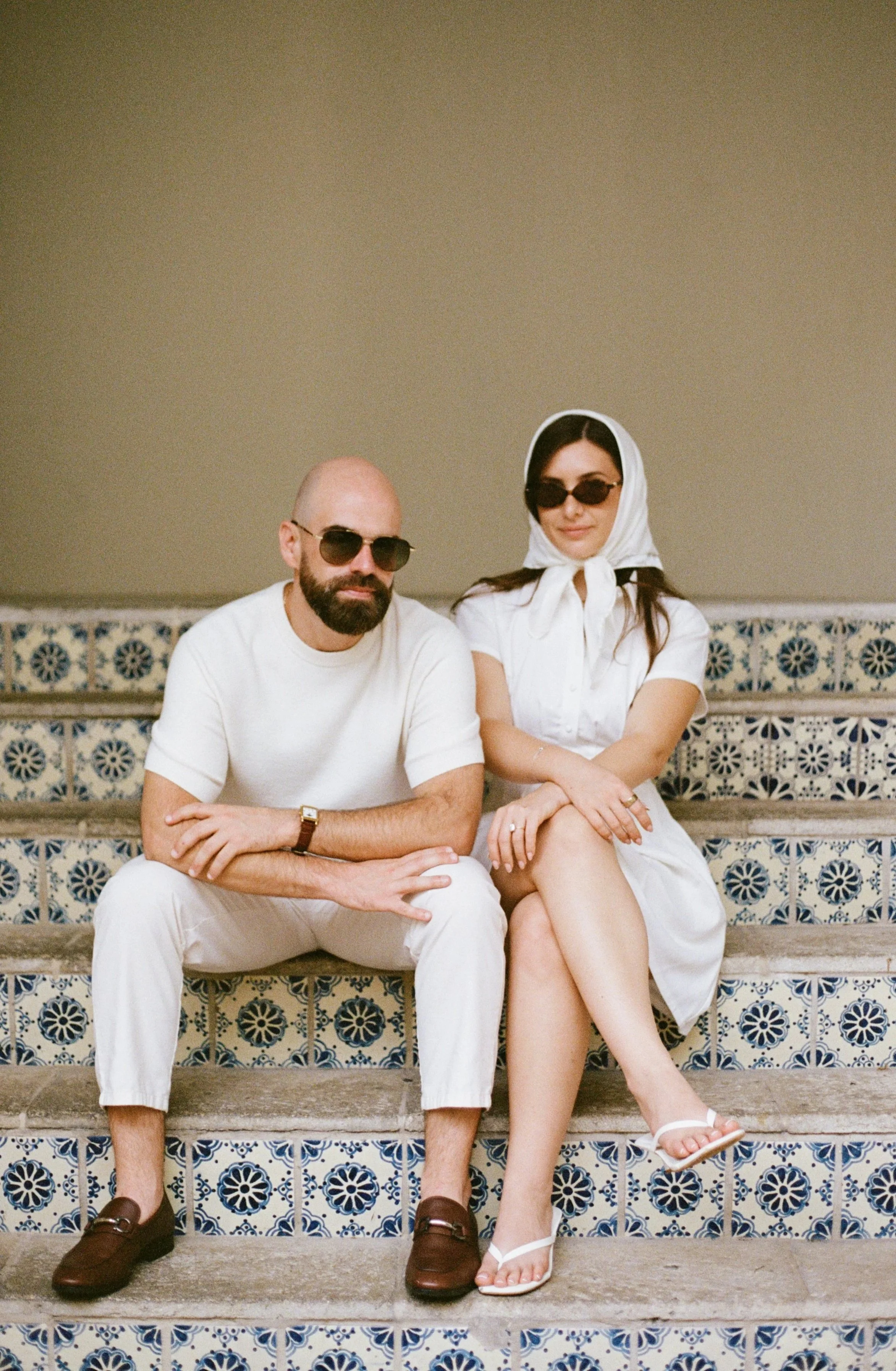 A man and woman sitting on decorative tiled steps, both wearing sunglasses and dressed in summer white clothing. The man has a beard, is bald, and wears a white t-shirt and white pants, with brown shoes and a watch. The woman has dark hair, is wearin