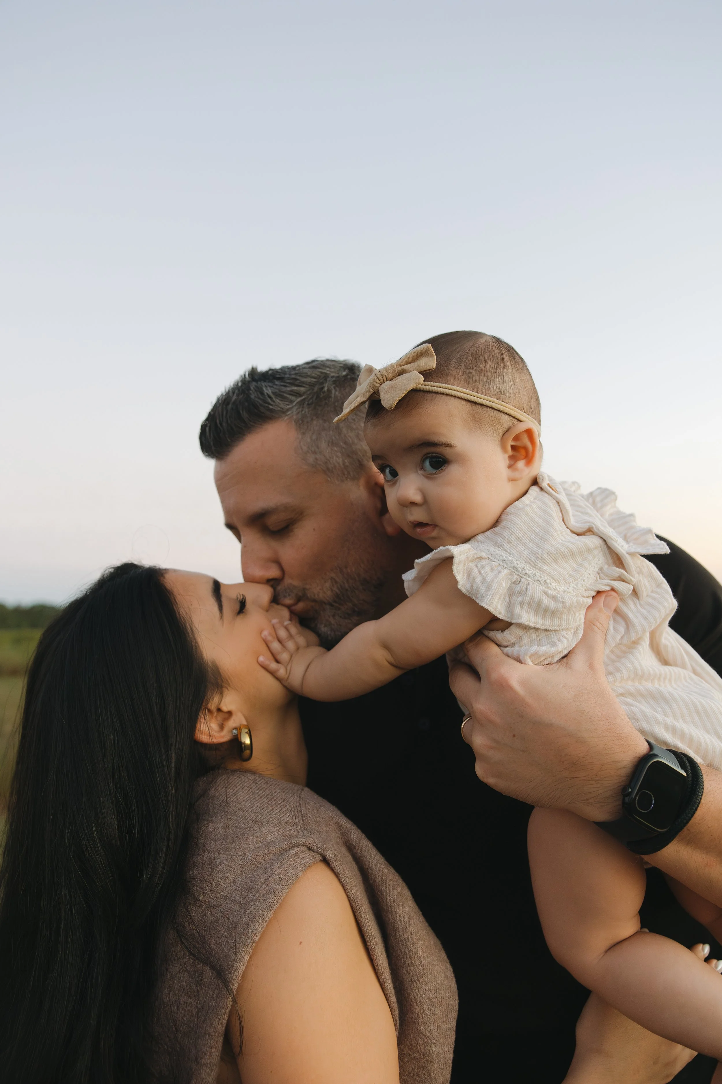 A man and woman kissing while holding a baby girl, outdoors during daytime. The woman has long black hair, and the man has short dark hair. The baby girl has a beige headband with a bow, blue eyes, and is wearing a light-colored dress with ruffles.