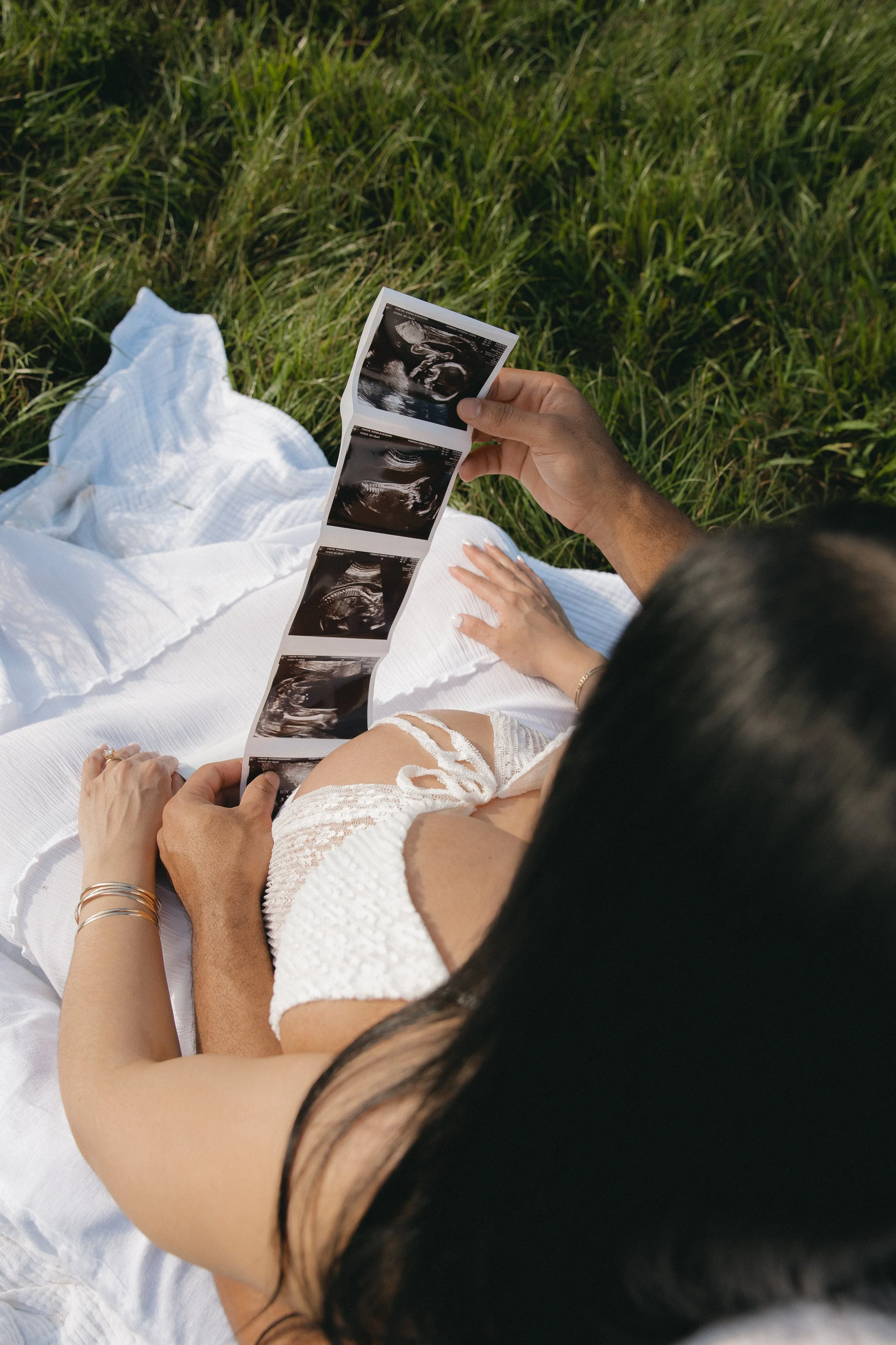 A person holding ultrasound images while sitting on a white blanket outdoors in a grassy area.