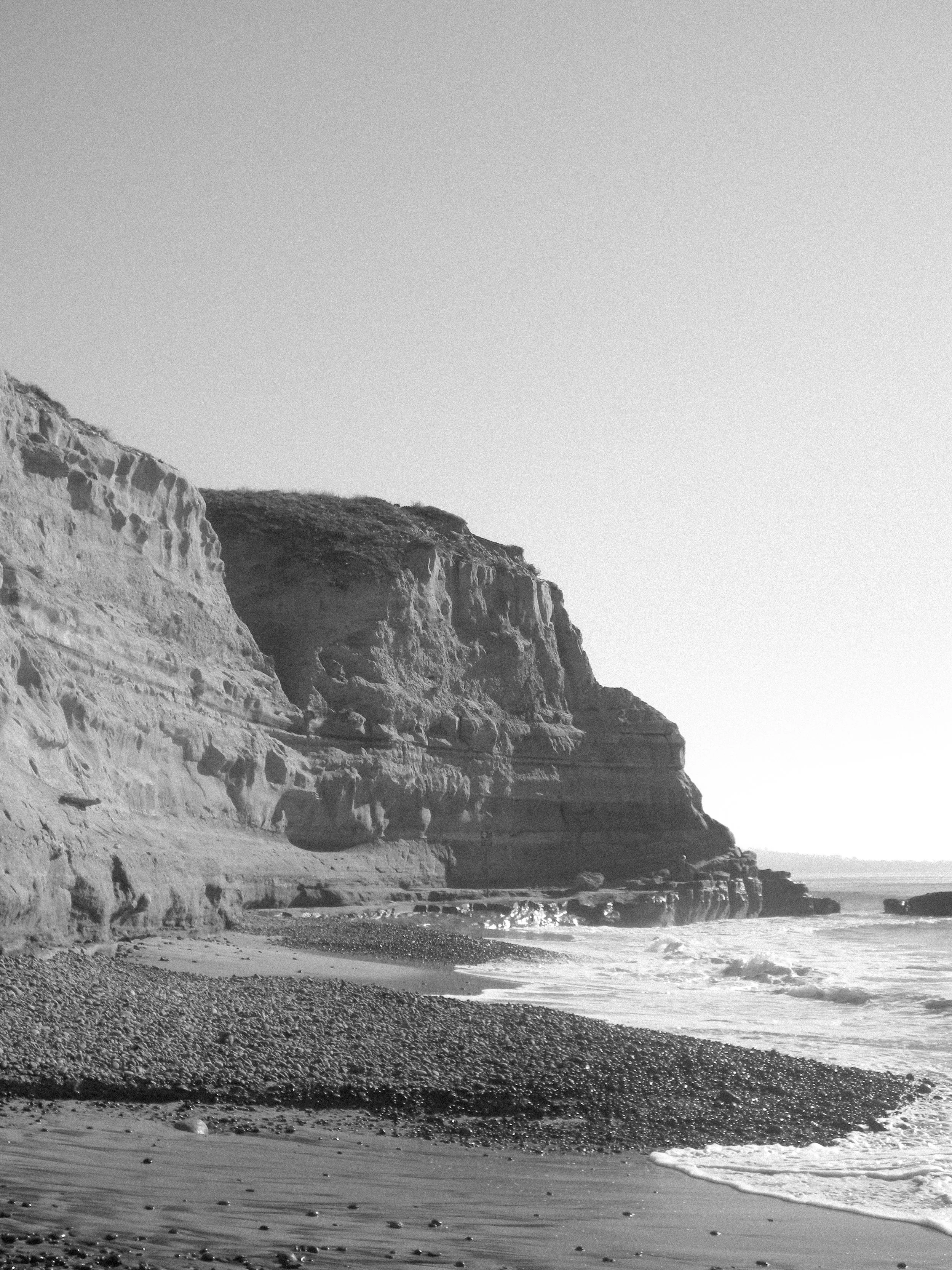 Black and white photo of a coastal cliff with the ocean at the base, small rocks on the beach, and a clear sky.