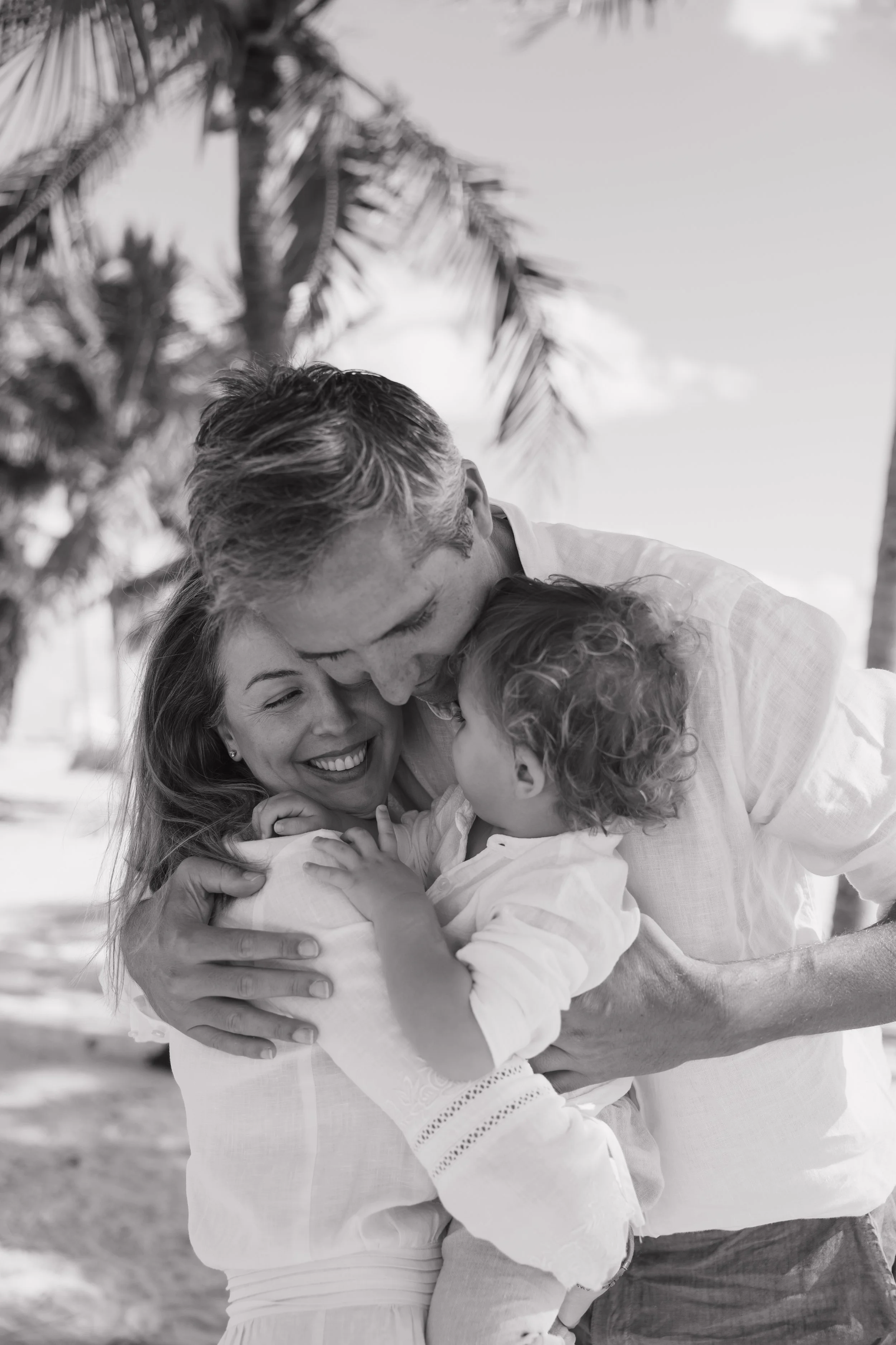 A family hugging and smiling outdoors, with palm trees in the background, in black and white.