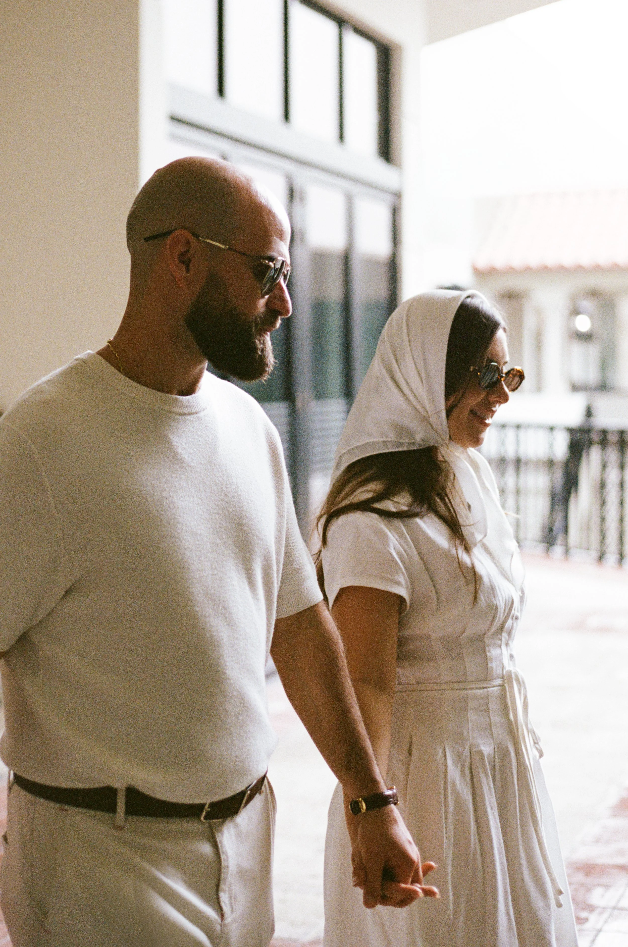 A man and a woman are holding hands, walking indoors near a large window with natural light. The man has a bald head, beard, sunglasses, and is dressed in a white shirt and beige pants. The woman is wearing a white hooded dress, sunglasses, and has l