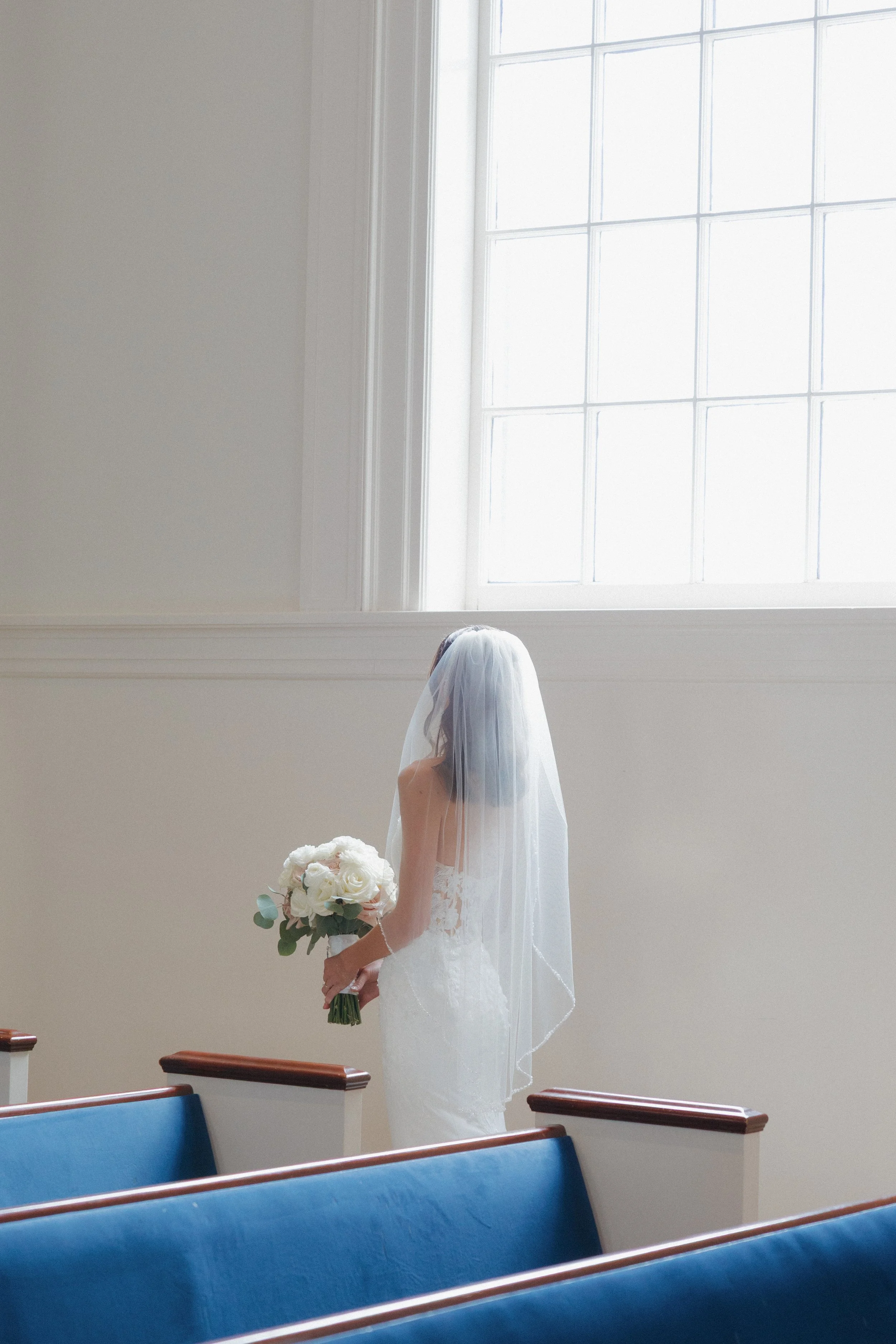 A bride in a white wedding dress and veil stands in a church, holding a bouquet of white flowers. She is near a large window with sunlight streaming through, and the pews are visible in the foreground.
