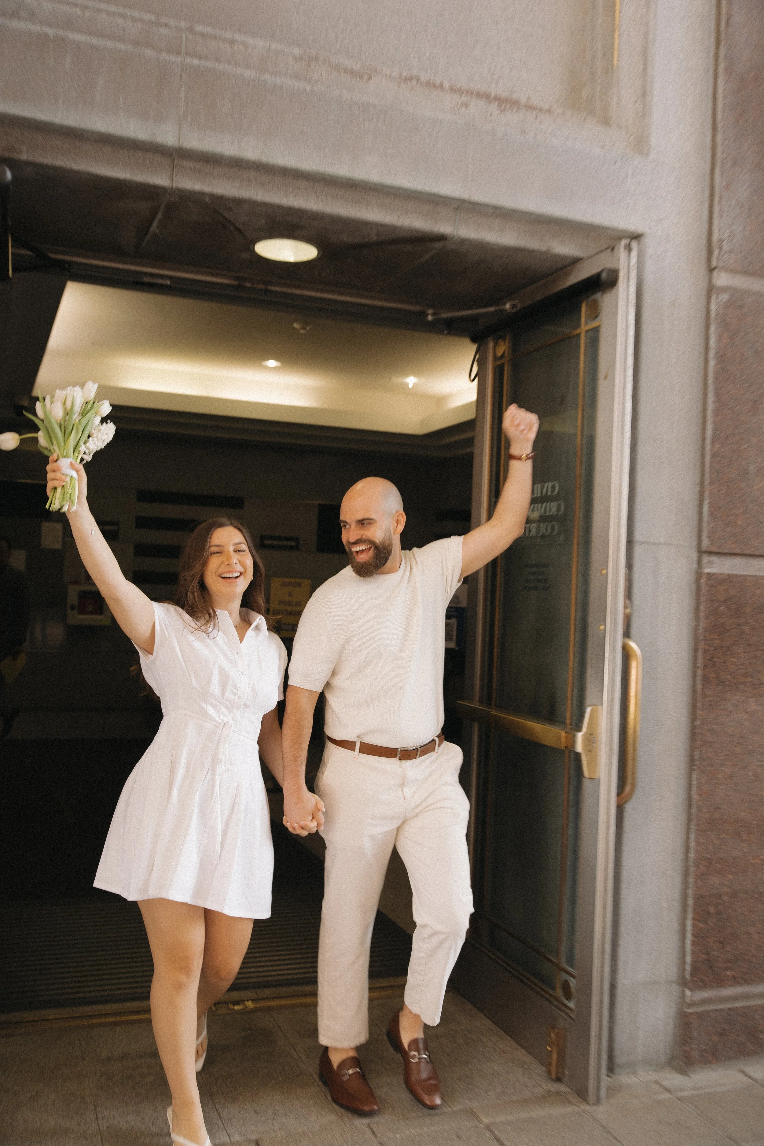 A smiling couple standing outside a building, holding hands, with the woman holding a bouquet of white flowers and the man raising his fist in celebration.