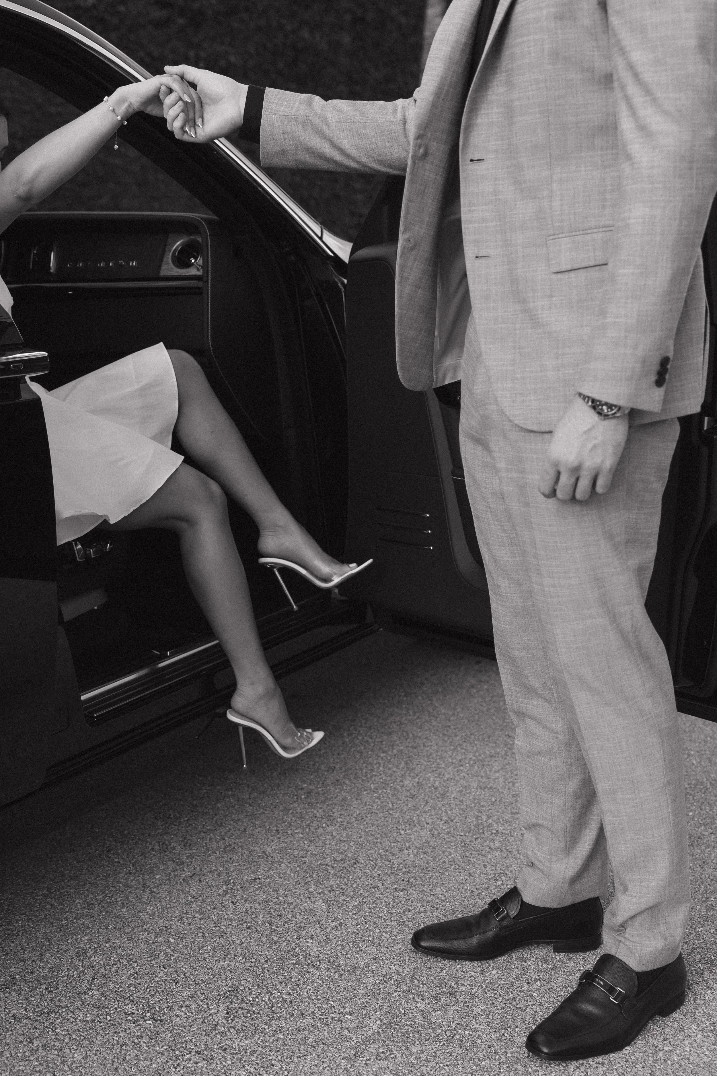 A man in a gray suit holding the hand of a woman sitting in a car. The woman is wearing a white dress and high-heeled shoes.
